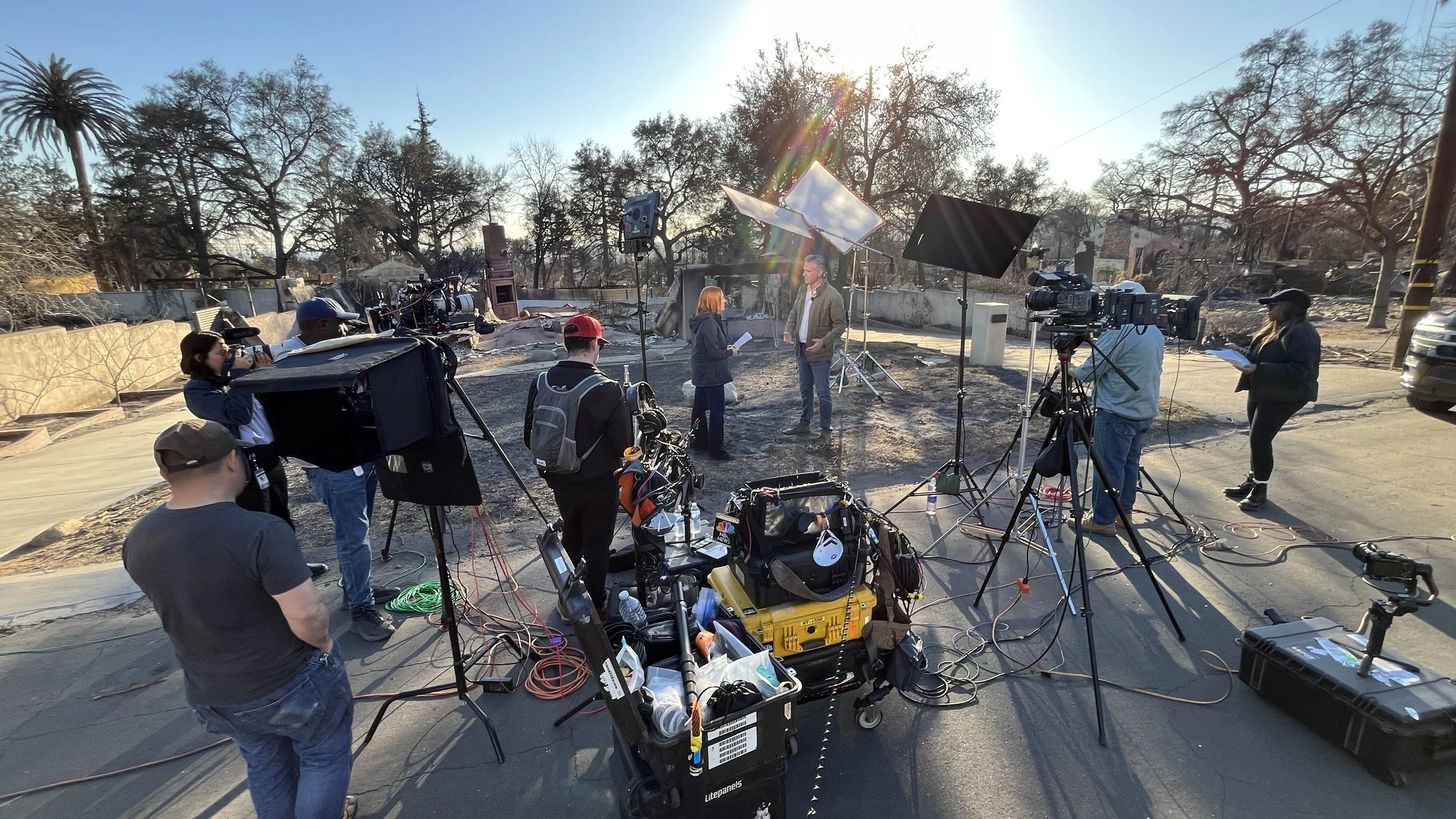 Filming crew setting up equipment outdoors with a man and woman speaking, in a yard with leafless trees and a fence, during sunlight.