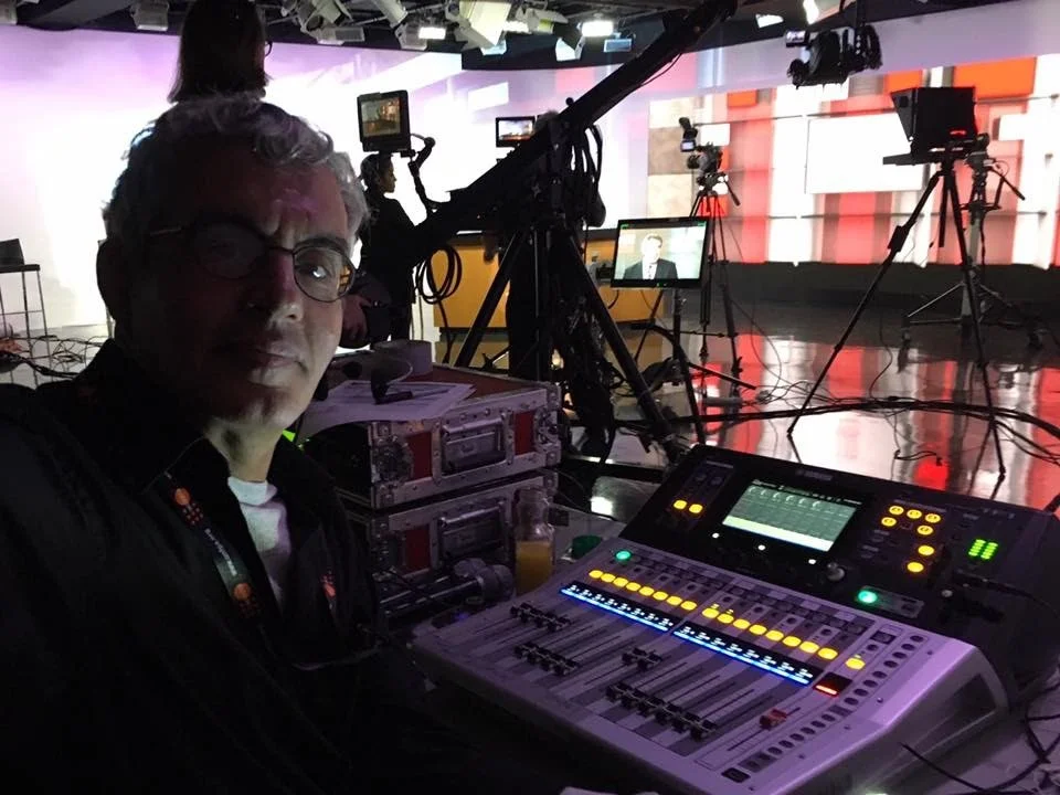 A behind-the-scenes view of a TV studio set with camera equipment, monitors, and lighting. A person is sitting at audio mixing equipment in the foreground.