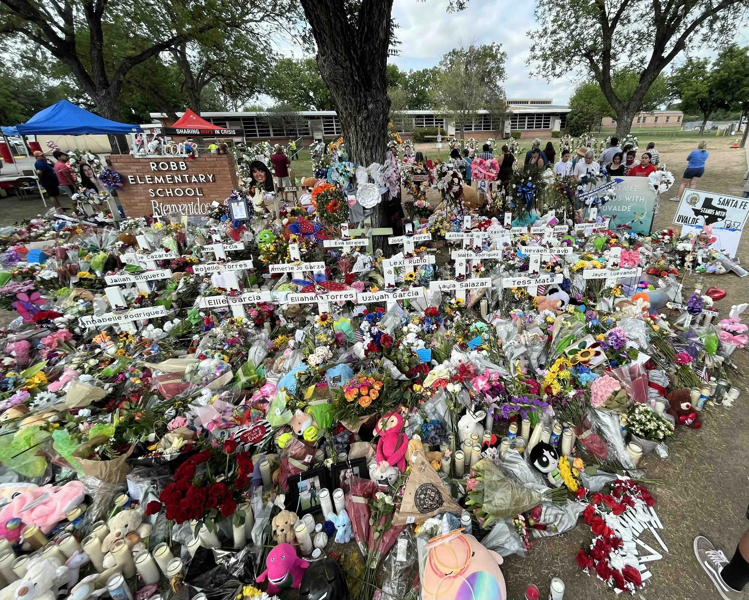 A large memorial with numerous flowers, stuffed animals, candles, and signs of tribute, set up in front of a school building, with children and adults gathered around.
