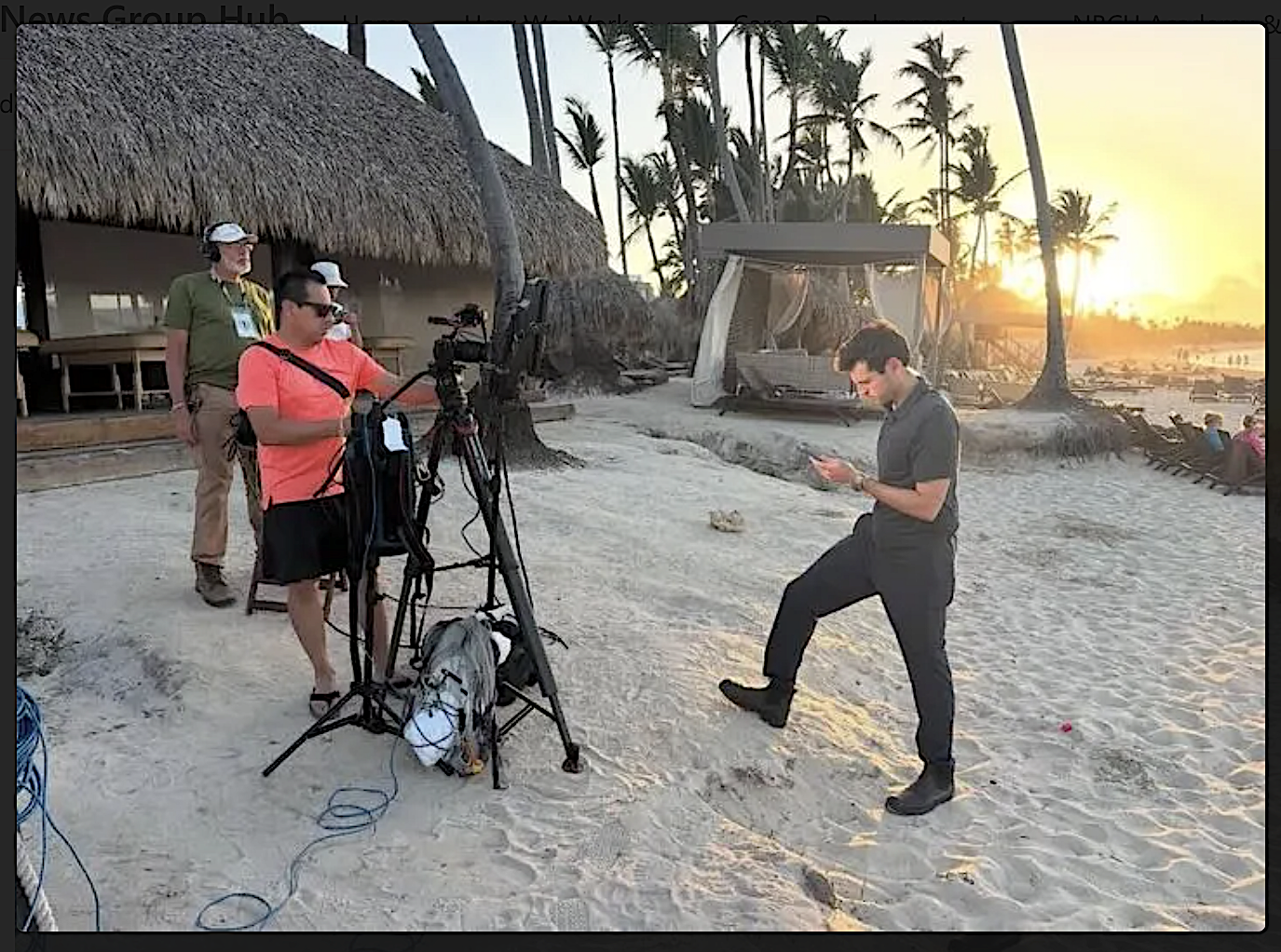 Filming crew setting up camera on a beach at sunset, with palm trees, wooden structures, and a person with a phone in the foreground.