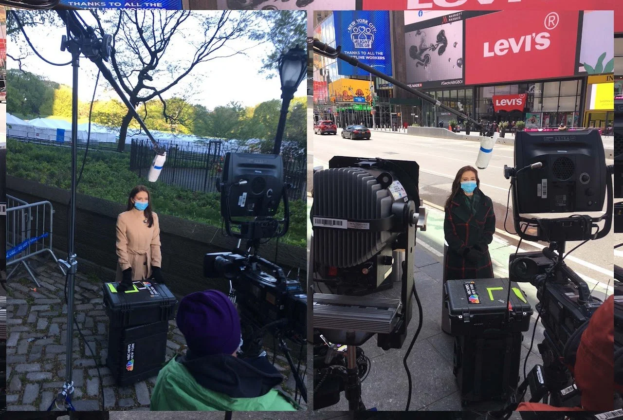 Two women wearing masks and standing outdoors in an urban area, surrounded by professional camera equipment. The woman on the left is dressed in a beige coat, and the woman on the right is in a dark coat with a plaid pattern. The background shows cit