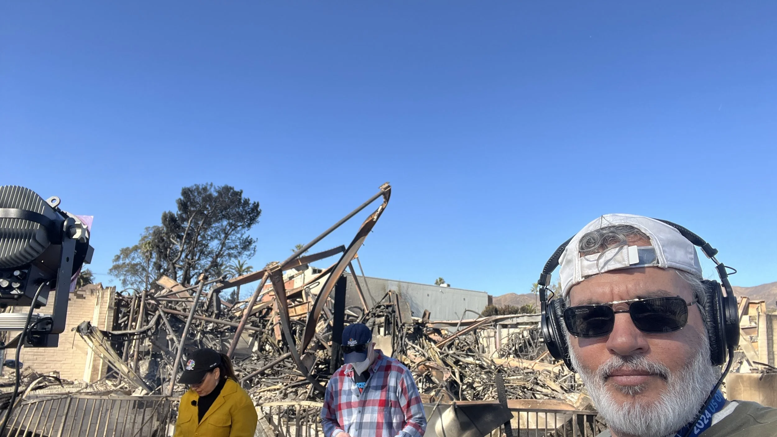 A man wearing sunglasses, a cap backwards, and headphones takes a selfie in front of a destroyed building with visible debris, twisted metal, and remains of walls. Two other people with face masks and casual clothing stand nearby.