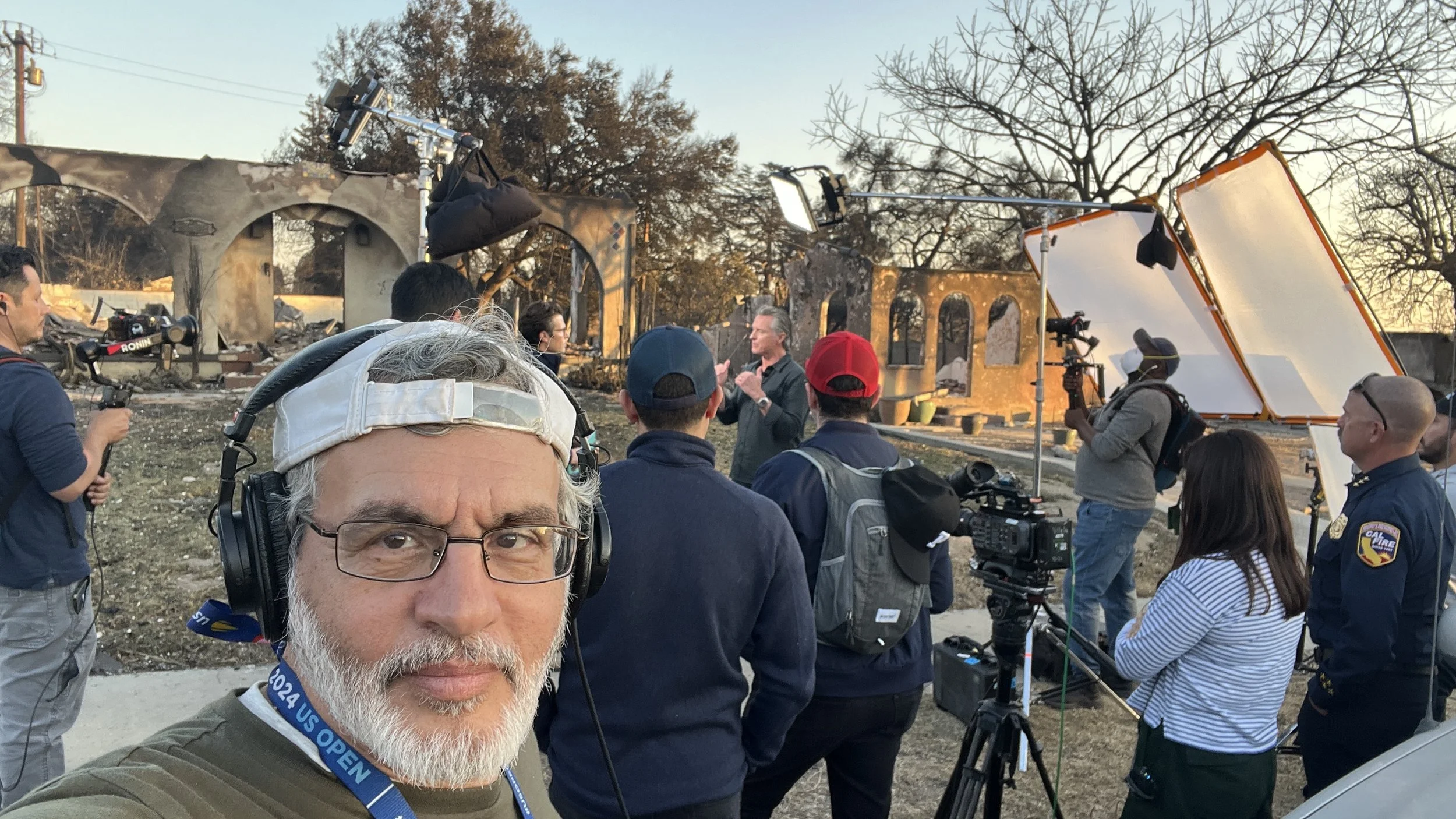 Group of people filming a news segment or documentary outdoors near a damaged building with smoke damage, with a man in the foreground wearing headphones and a lanyard, and a person in the background recording and controlling equipment.