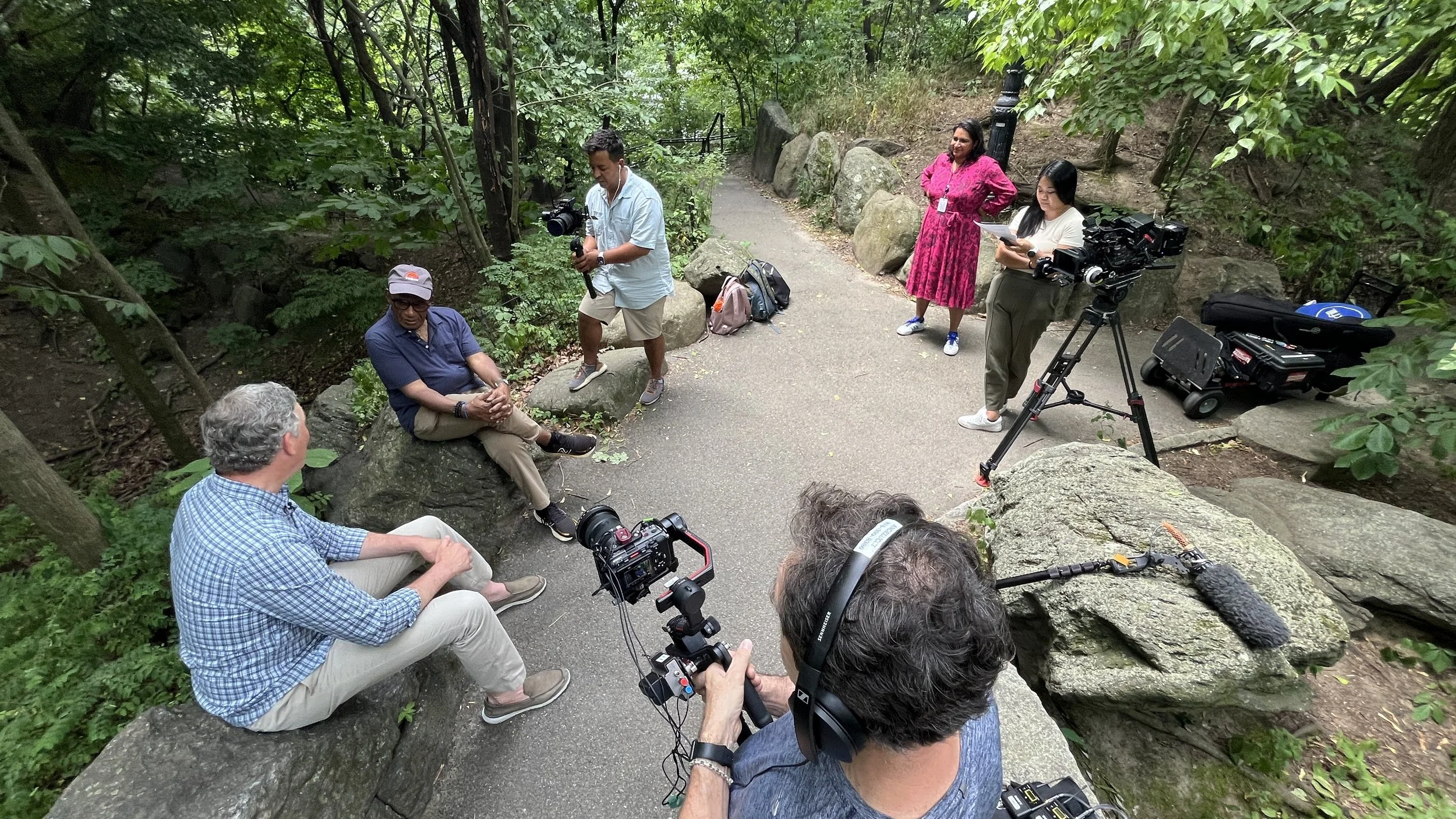 A news crew interviews a man on a park trail surrounded by trees, with cameras and equipment set up.