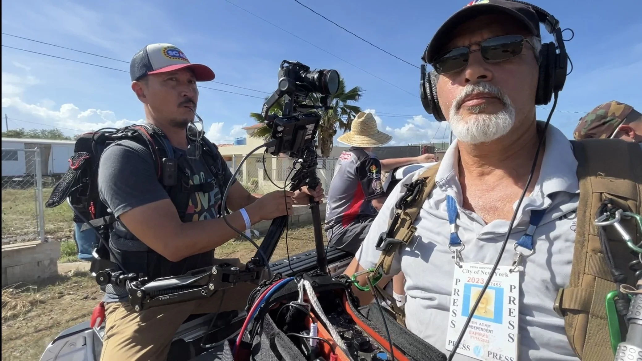 Two male journalists, one with a camera on a stabilizer and the other wearing headphones, outdoors in sunny weather with a blue sky and palm trees in the background.