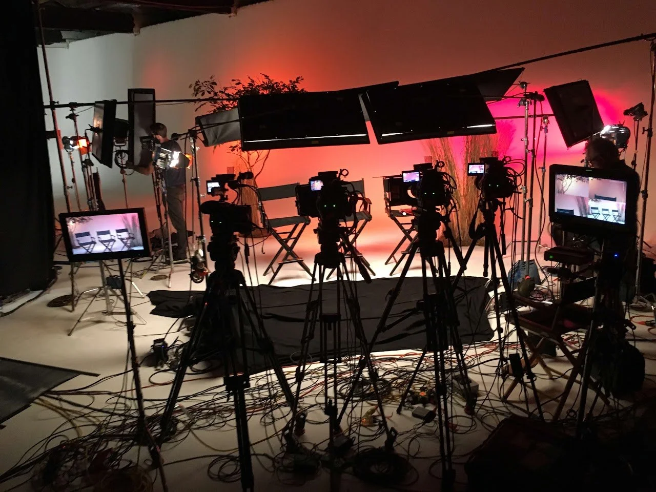 Television studio set with multiple cameras, monitors, lighting equipment, and a backdrop with colored lighting.