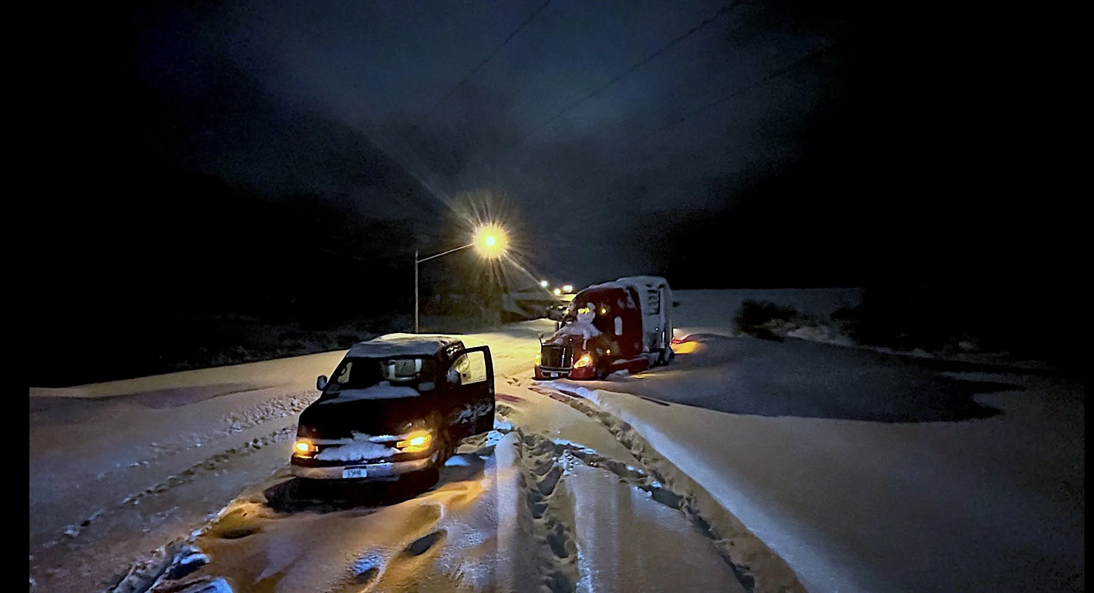 Nighttime snowy road with two parked vehicles, a small black car in the foreground and a larger semi-truck in the background, illuminated by a streetlight.