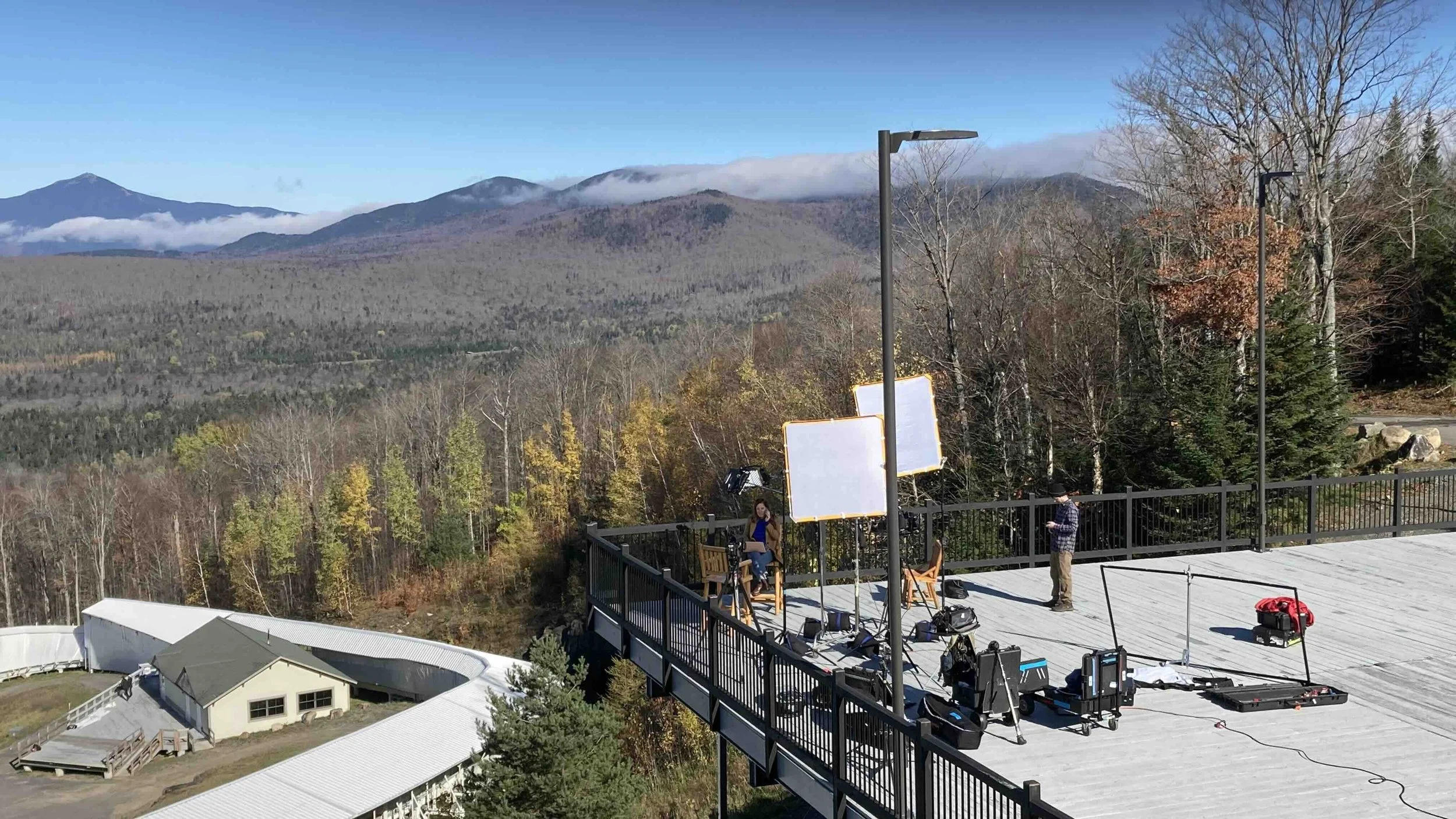 A rooftop overlooking a forested mountain landscape with a distant snow-capped peak, and a filming crew setting up equipment on the rooftop.