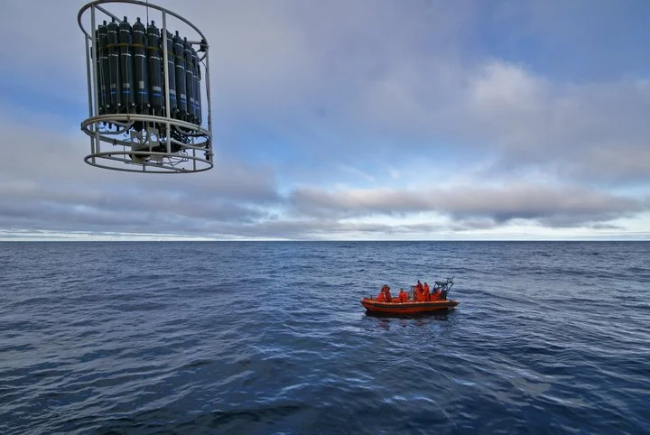 Sea with a drone carrying multiple cylinders and a boat with people in orange suits.