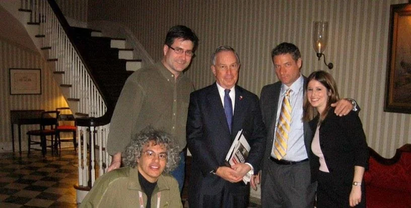 Group of five people posing in a room with striped wallpaper, staircase, and framed pictures, smiling at the camera.
