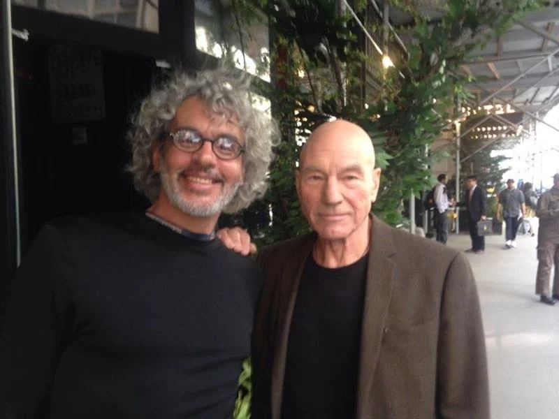 Two men standing next to each other outdoors, one with curly gray hair and glasses, the other with a shaved head and wearing a blazer, with a city street and people in the background.