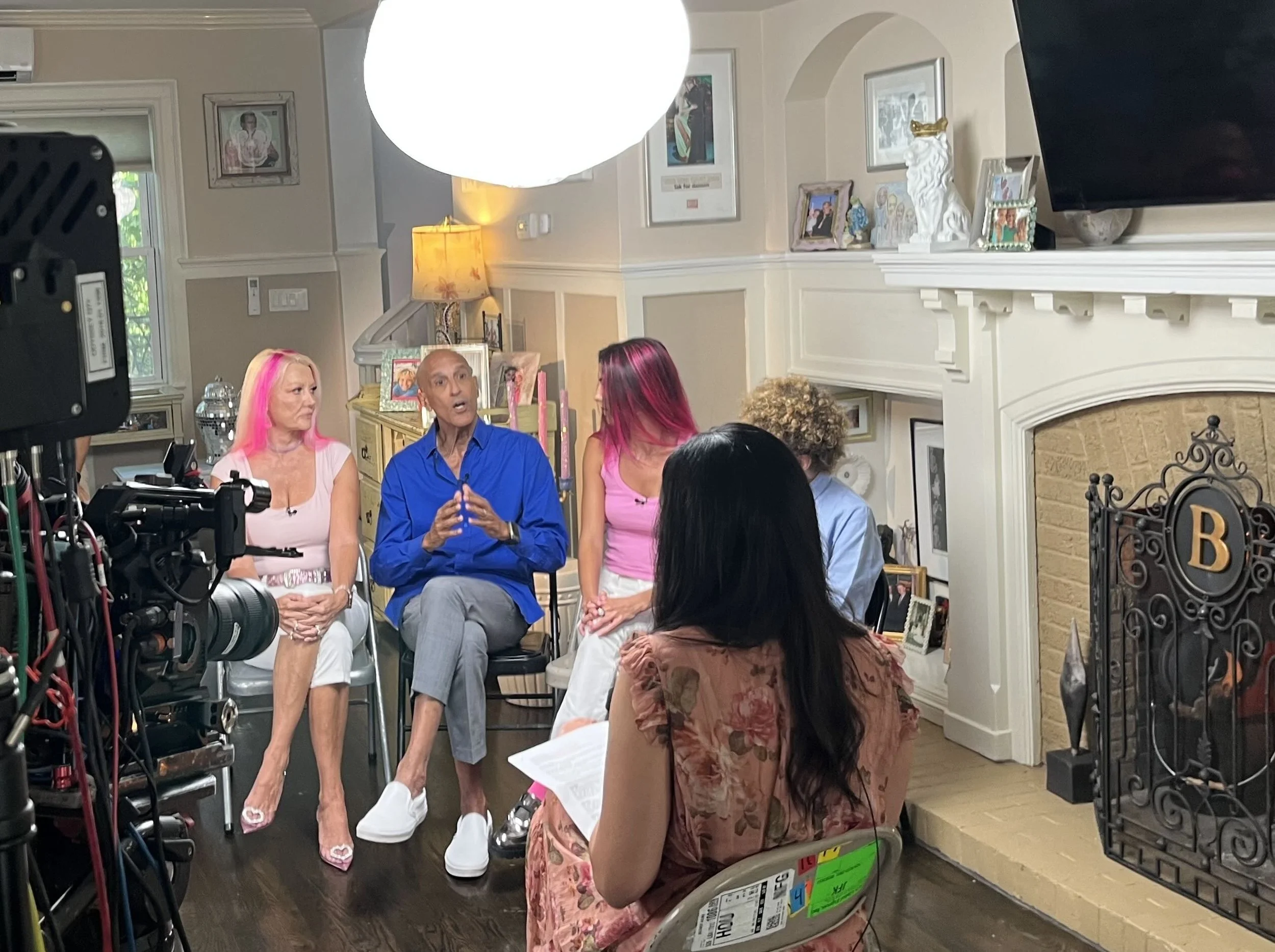 Group of people sitting in a living room during a recording or interview, with a camera and lighting equipment visible, and a woman in the foreground holding papers