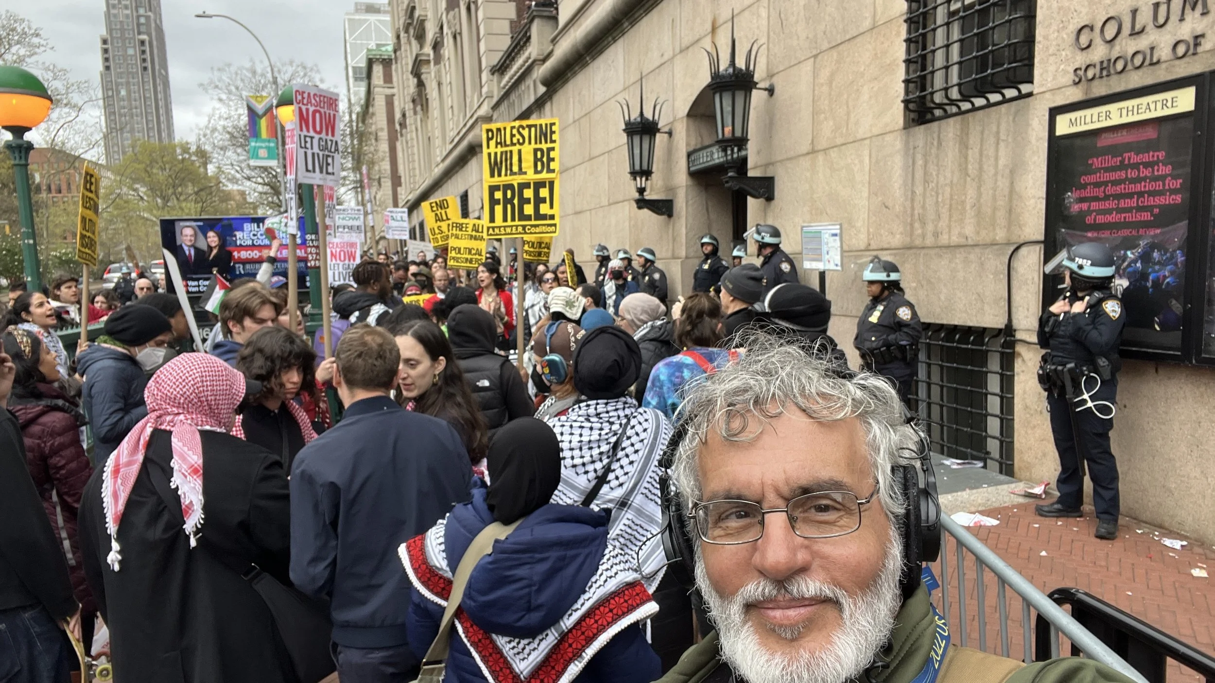 A diverse group of protesters gathered outside a building with police officers standing guard. Protesters hold signs with messages such as 'Palestine will be free' and 'Ceasefire now, let Gaza live.' The man in the foreground is taking a selfie, wear