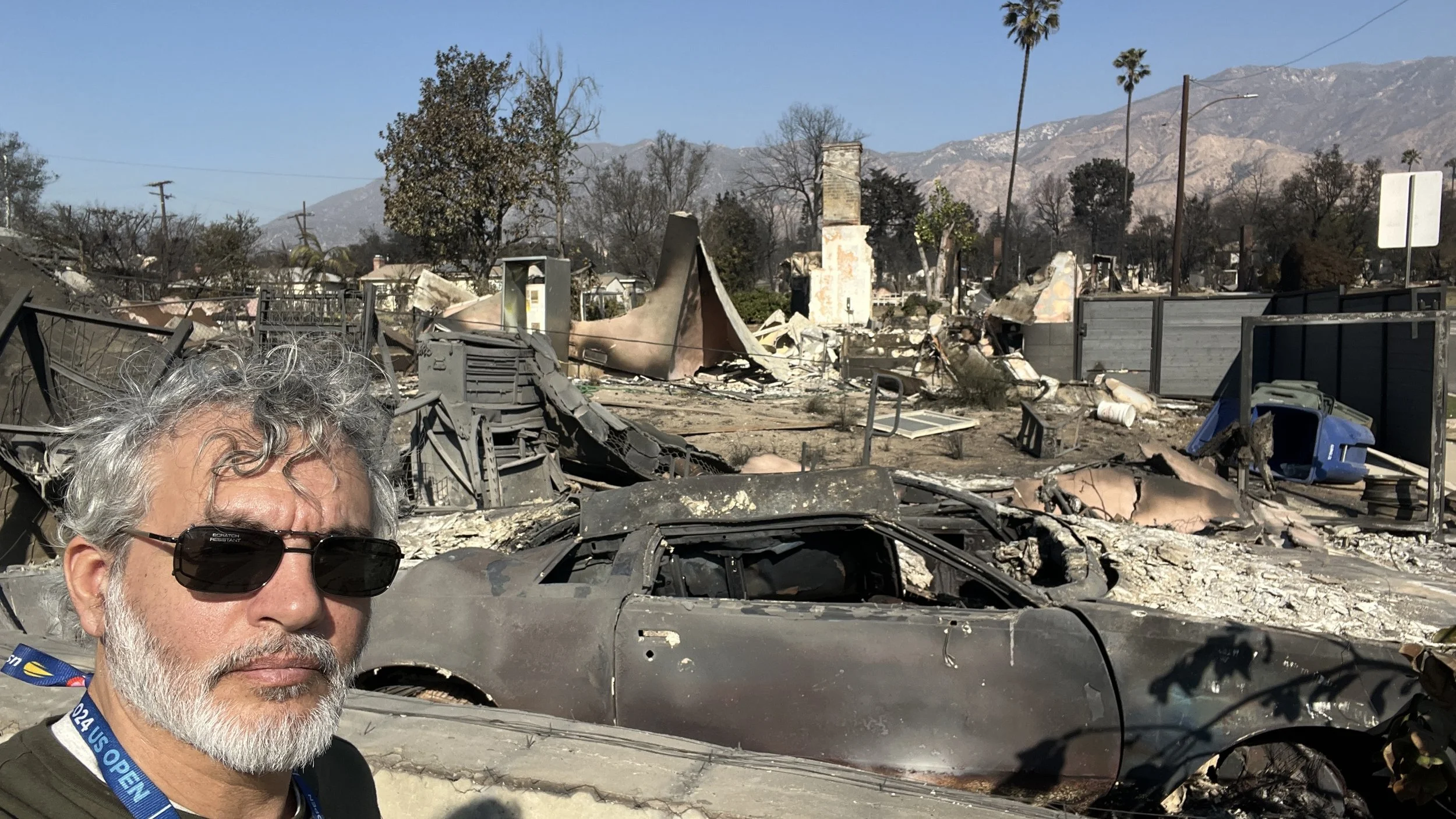A man with gray hair, beard, sunglasses, and a blue lanyard takes a selfie at a site of destruction caused by a fire, with burned debris and a wrecked car in the background.