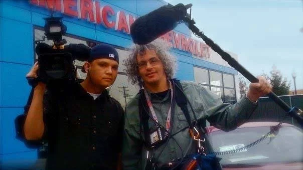 Two men standing outside a building with a blue exterior, one holding a camera mounted on a shoulder rig, the other holding a boom microphone, likely filming or recording.