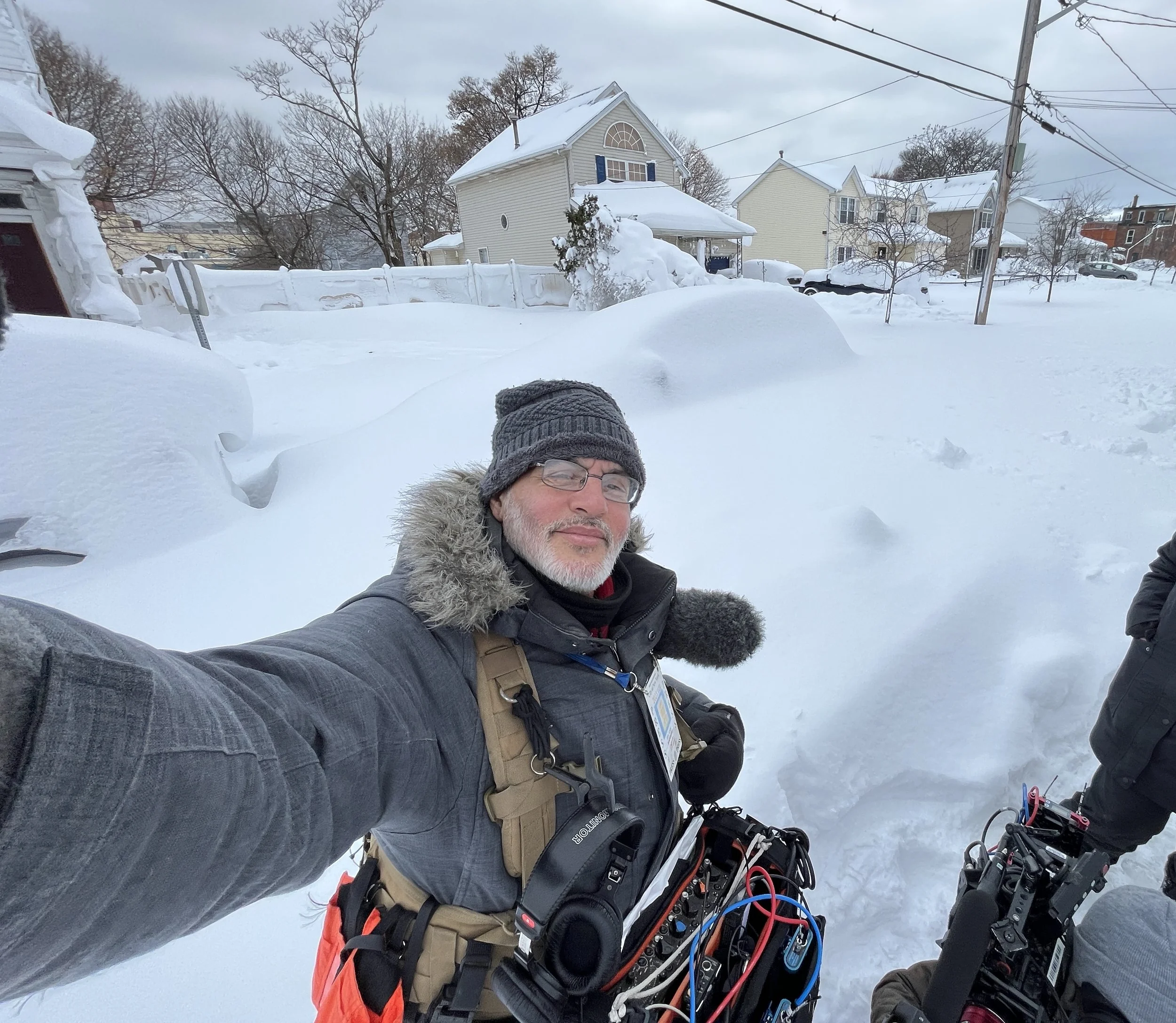 A man with a gray beanie, glasses, and a winter coat taking a selfie in a snowy yard with snow-covered houses and trees in the background.
