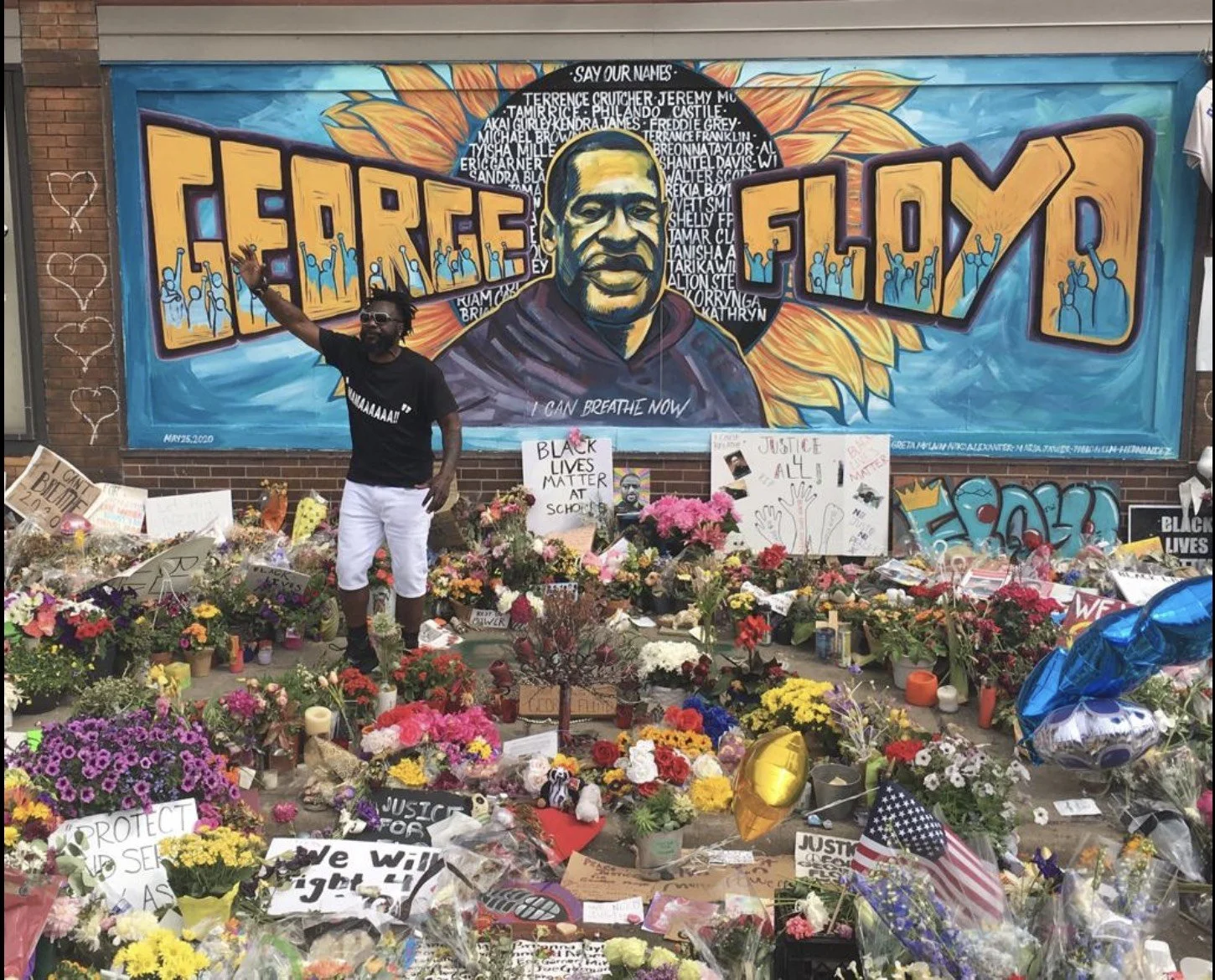 A memorial with numerous flowers, signs, and tributes in front of a large mural of George Floyd. The mural depicts George Floyd with a sunflower in the background, and the text 'George Floyd' is prominently displayed. A man stands among the tributes,