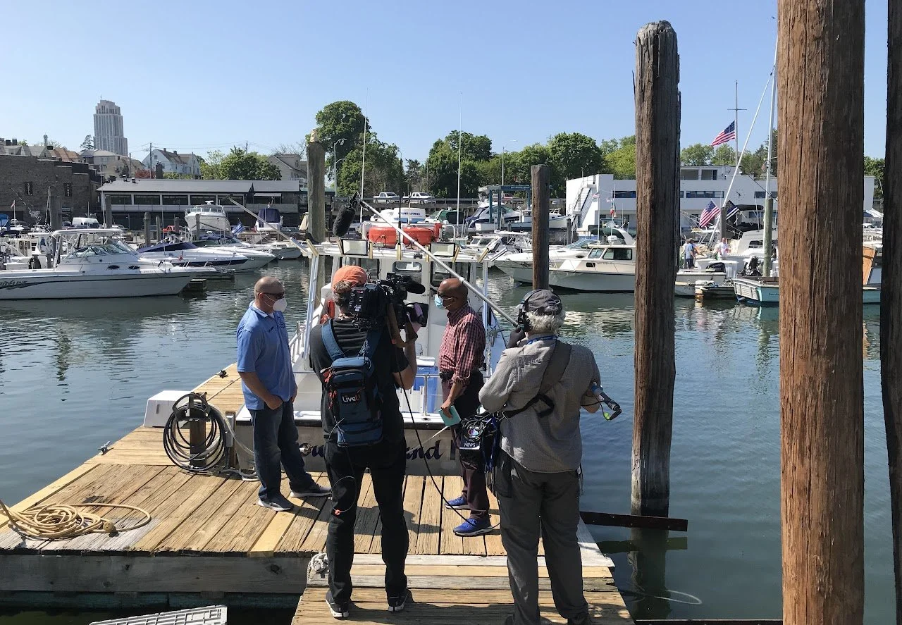 Group of people filming or interviewing on a dock at a marina, with boats and yachts in the background.