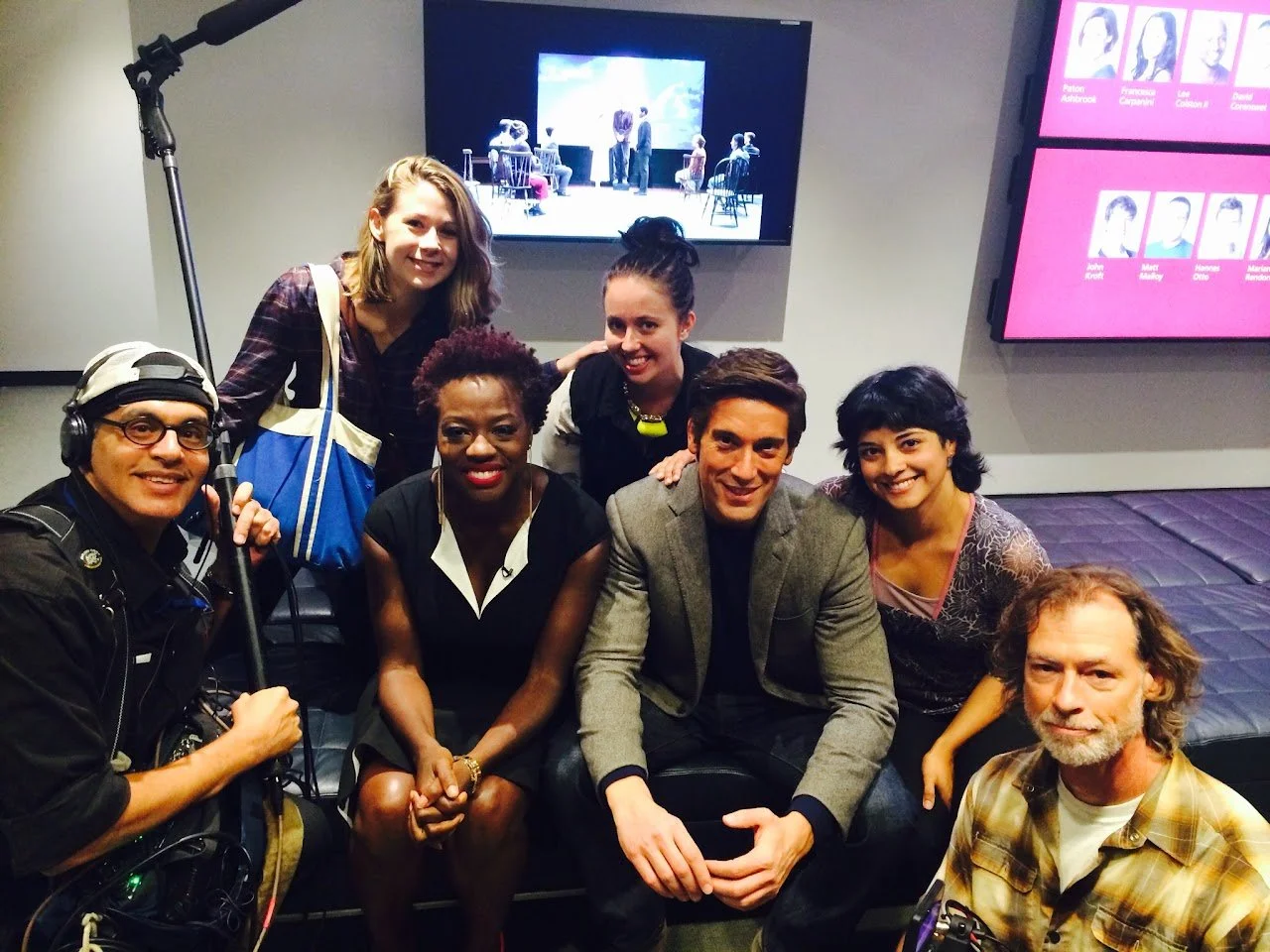 Group of eight people, including men and women, smiling and posing together in front of a monitor and multimedia screens, with professional film equipment visible.