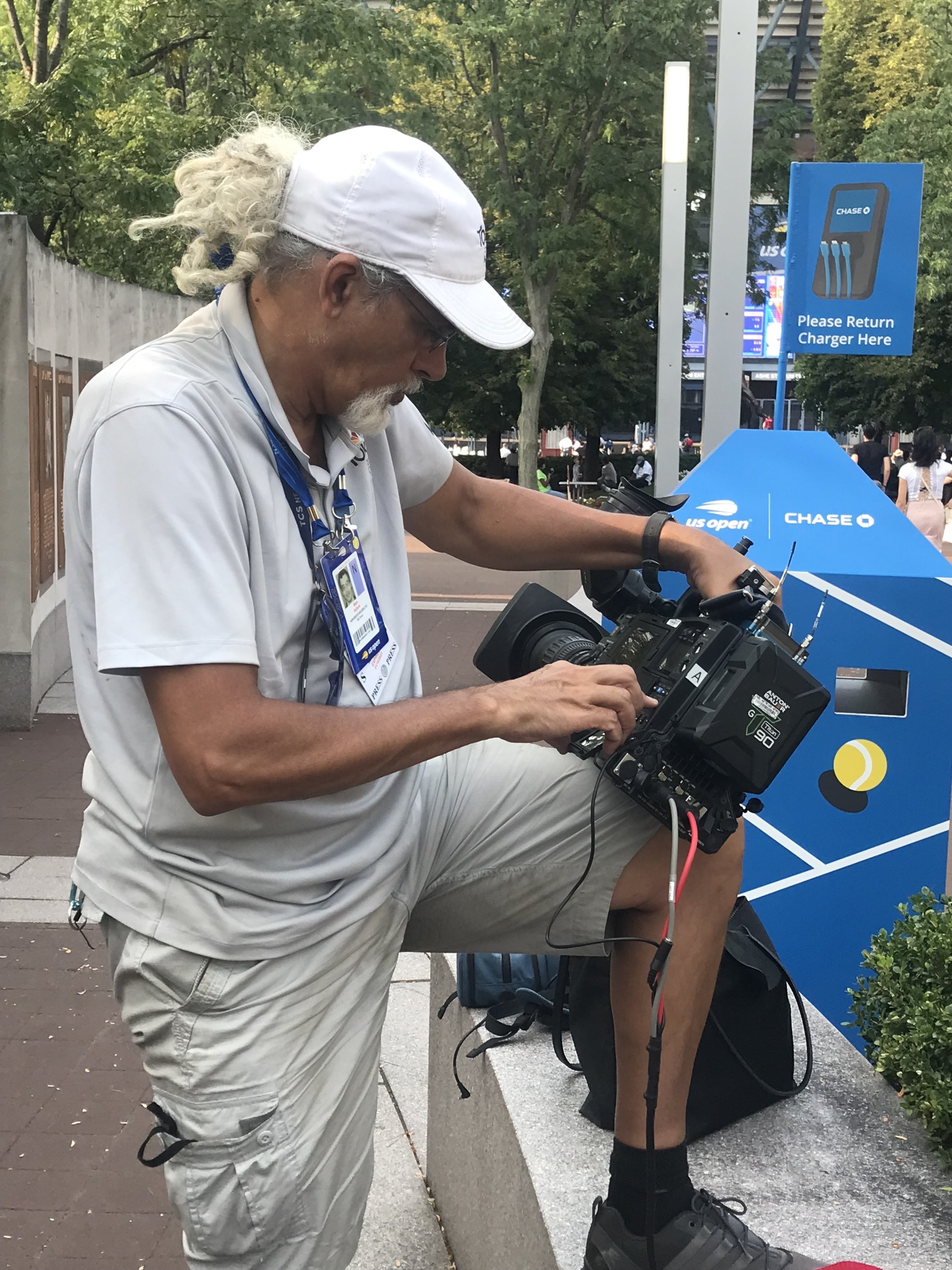A man with long gray hair, a beard, wearing a white cap, glasses, white polo shirt, and beige shorts, is sitting on a bench and operating a professional video camera.