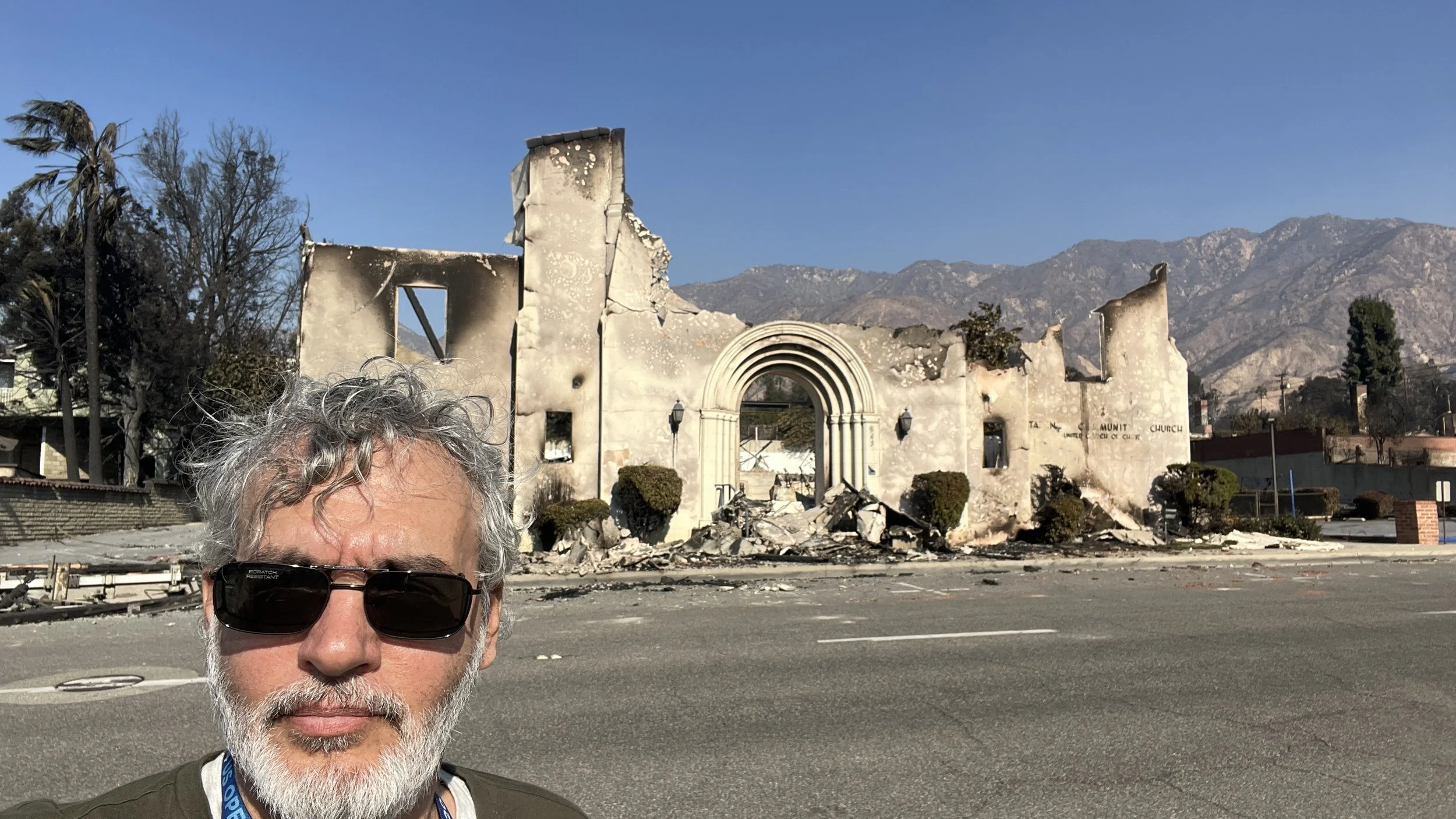 A man with gray curly hair and a beard wearing sunglasses in front of a burned and damaged church with a mountain range in the background.
