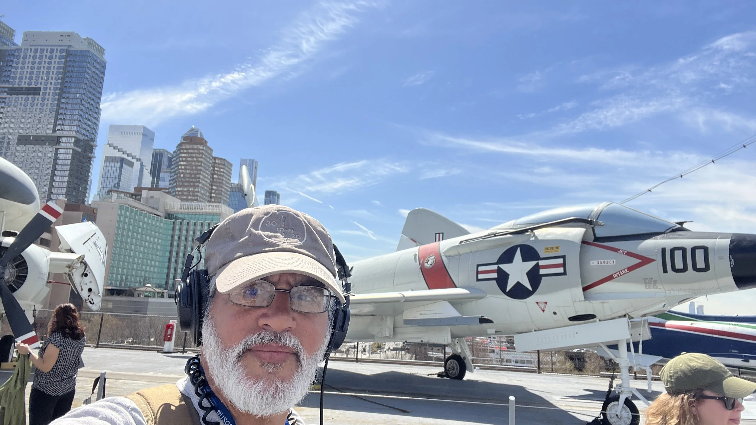 A man with glasses, a beard, and headphones takes a selfie at an outdoor aircraft exhibit in front of a jet fighter with a city skyline and tall buildings in the background on a sunny day.