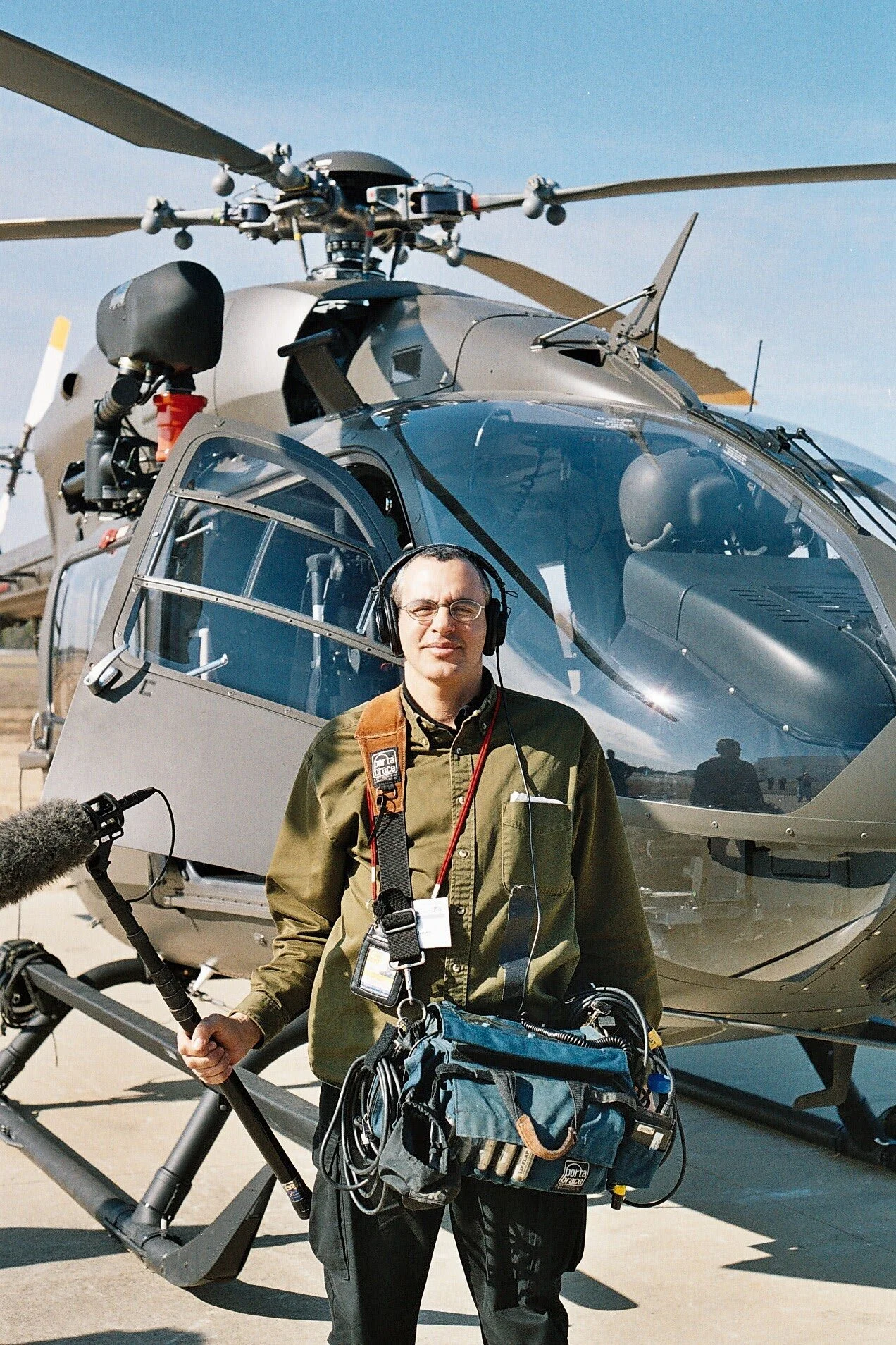 A man wearing glasses, headphones, and a green jacket stands in front of a helicopter, holding a boom microphone with a windscreen, and carrying a camera bag with equipment.