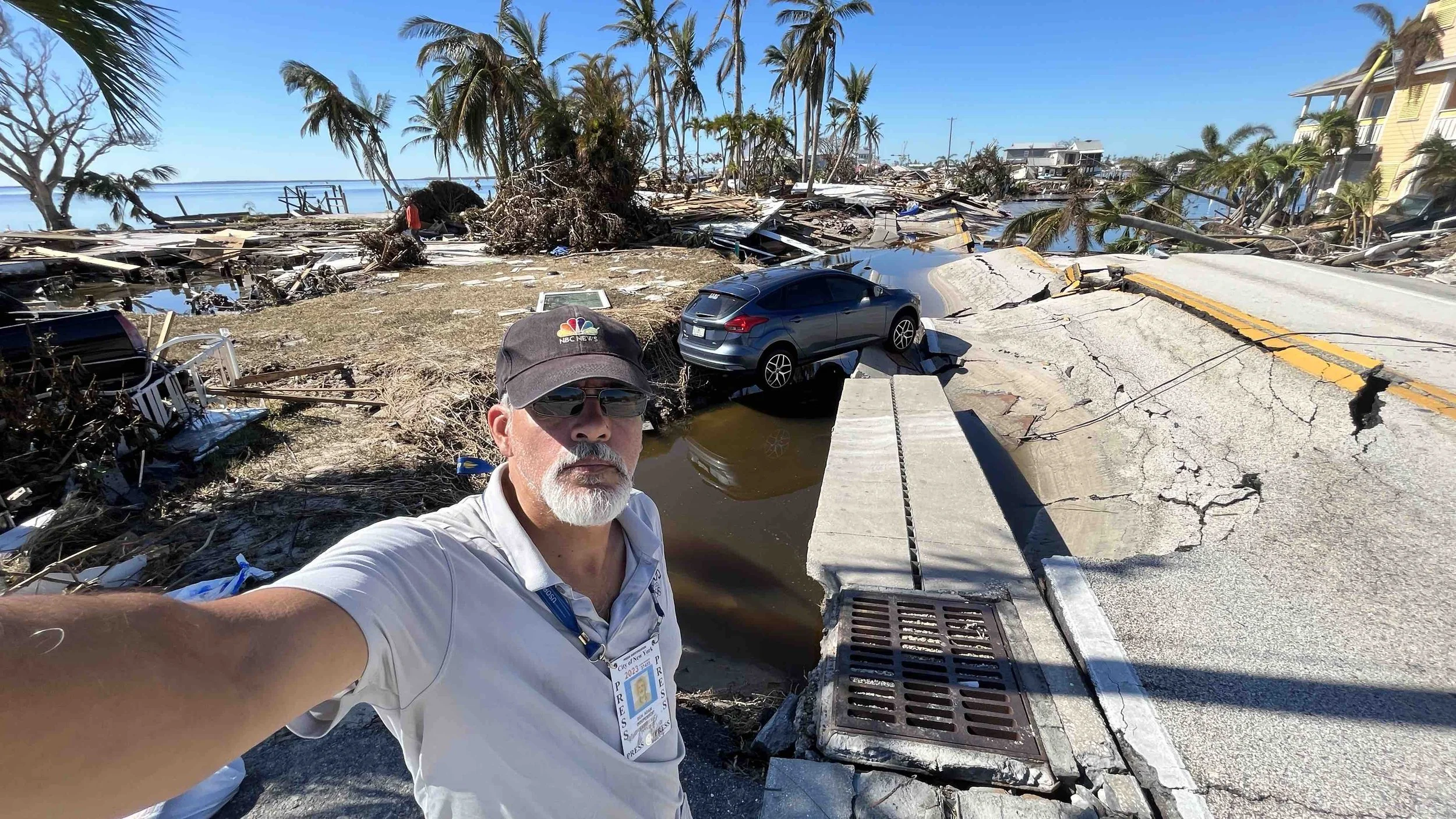 A man with a white beard taking a selfie at a destroyed coastal area after a hurricane or natural disaster, with damaged buildings, fallen trees, and a car overturned in the background.