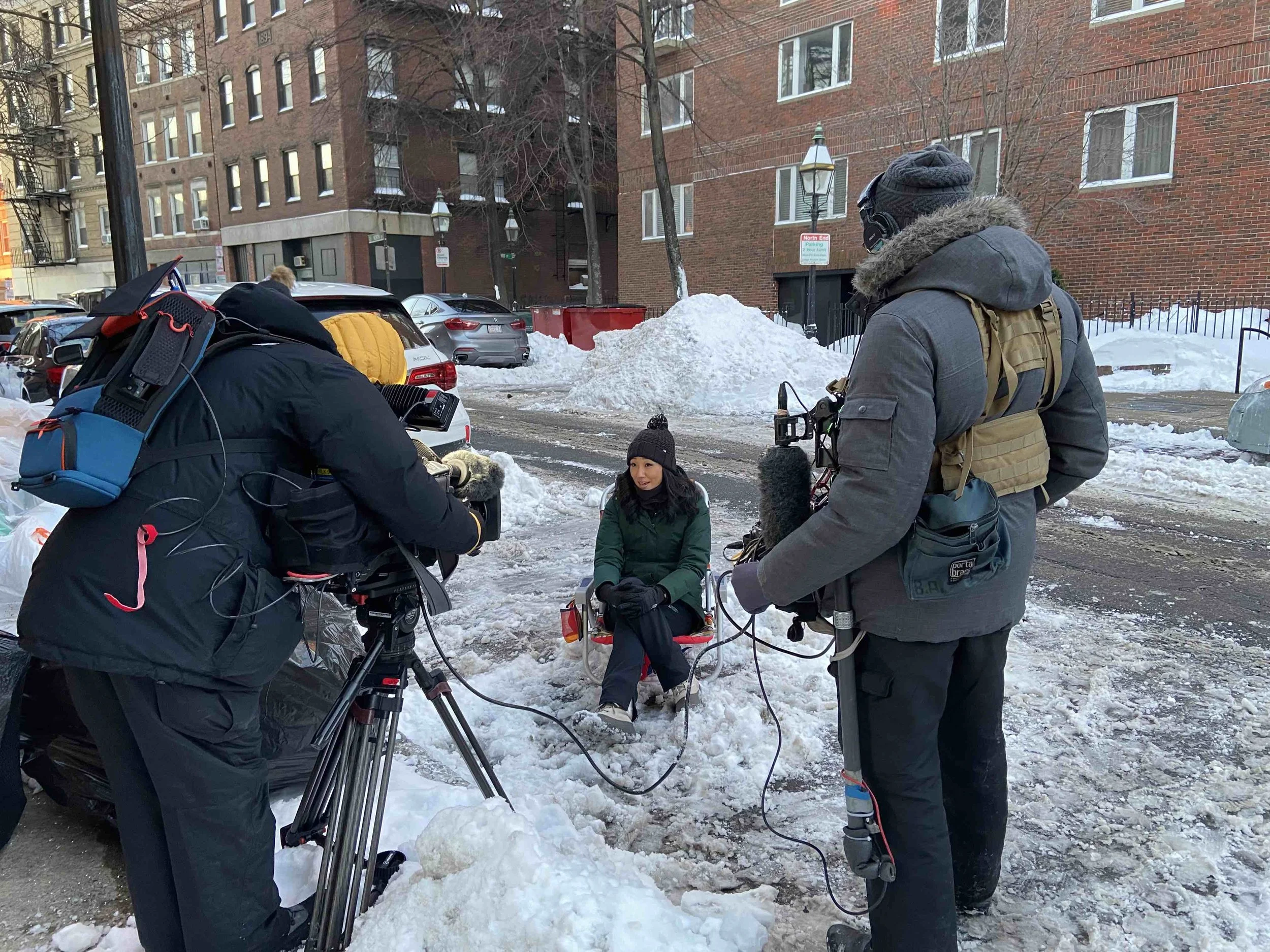 A woman being interviewed outdoors in winter, seated in a chair, while two cameramen film her. Snow is on the ground and on parked cars, with apartment buildings in the background.