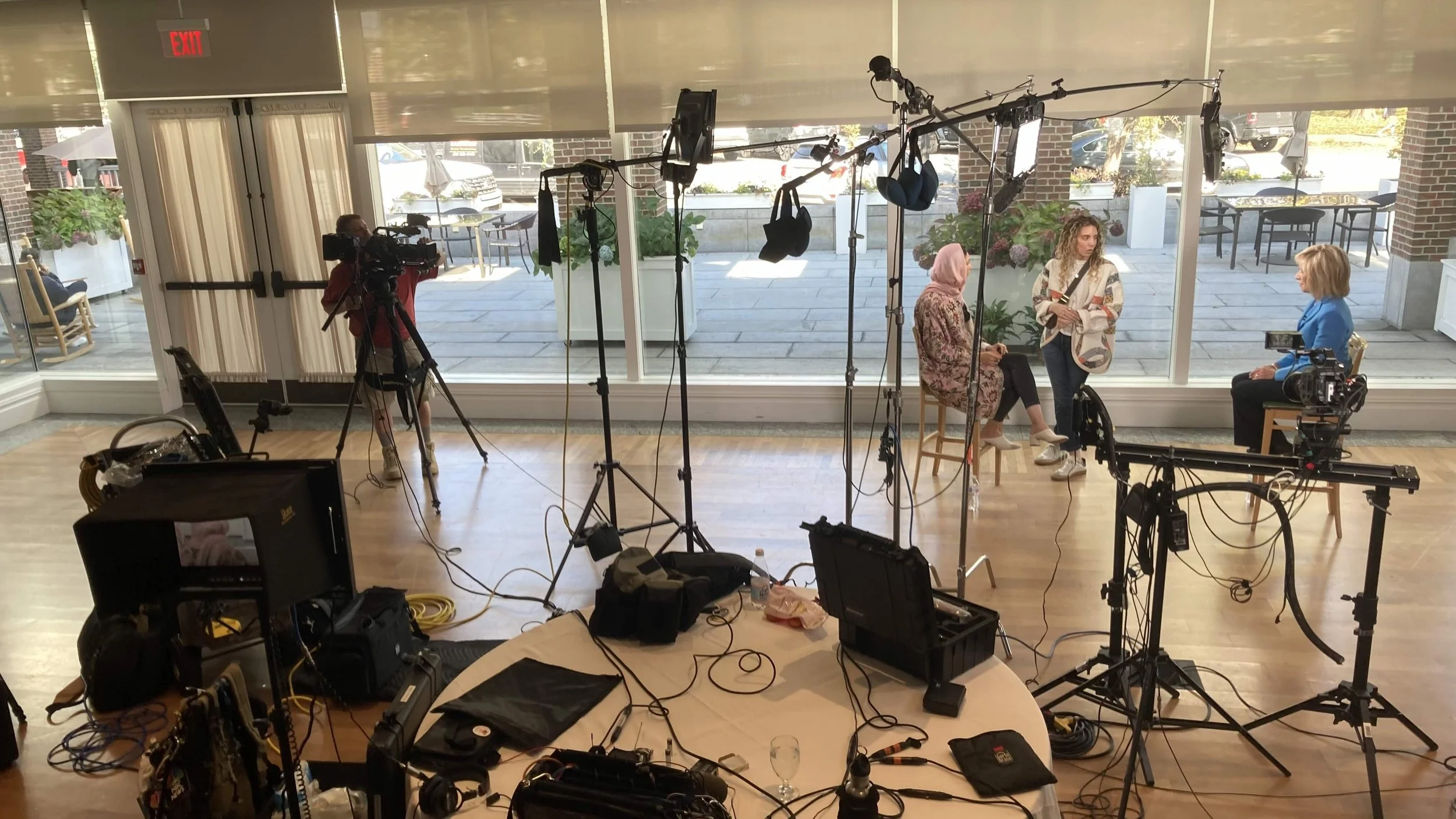 A filmed interview with three women in a well-lit room with large windows, cameras, lights, and other filming equipment visible in the setup.