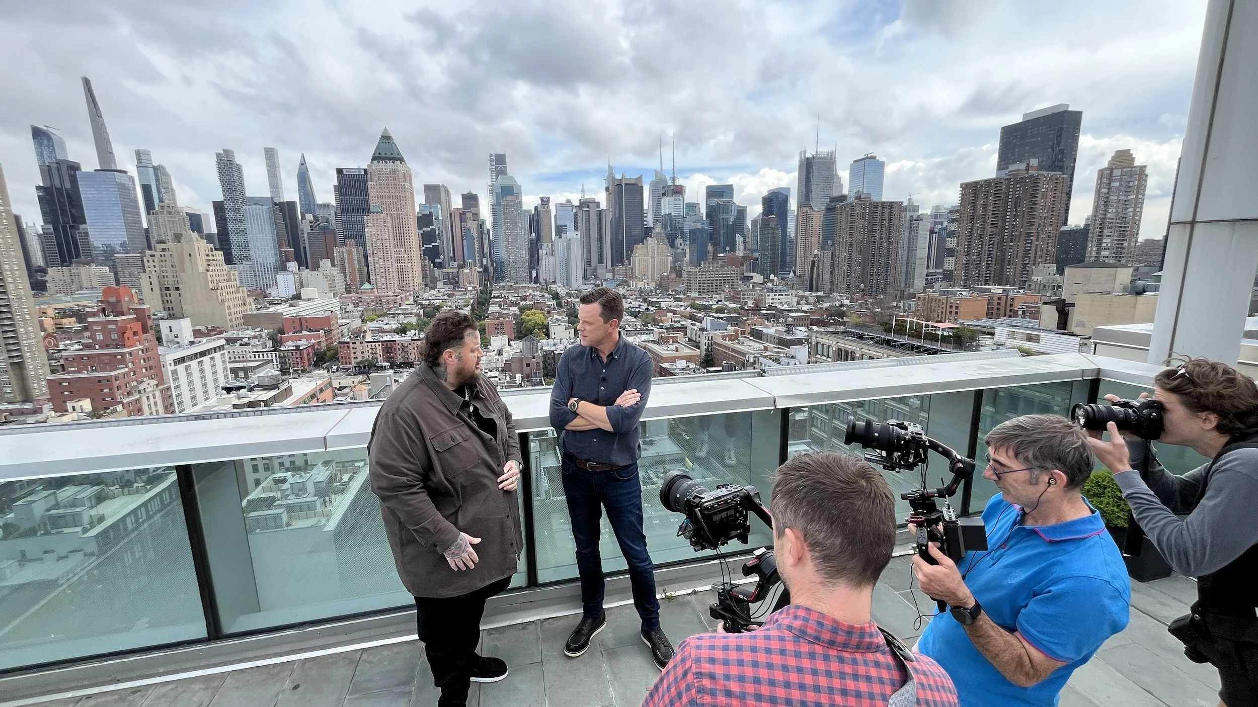 Two men are standing and talking on a rooftop with a city skyline in the background. Four people, including one using a camera, are filming them.