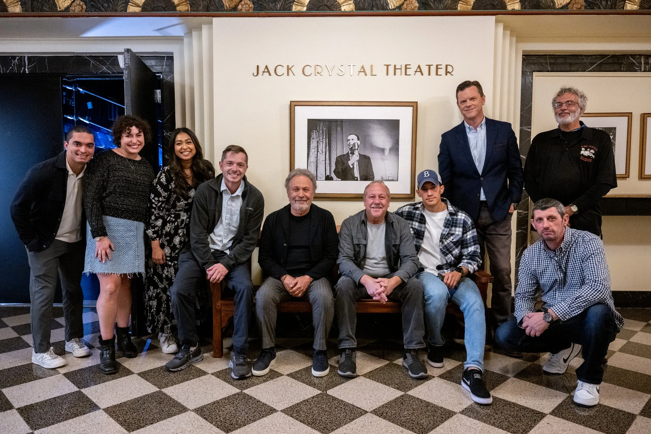 Group of eleven people sitting and standing in front of a wall with art and the text 'Jack Crystal Theater' at a theater lobby.