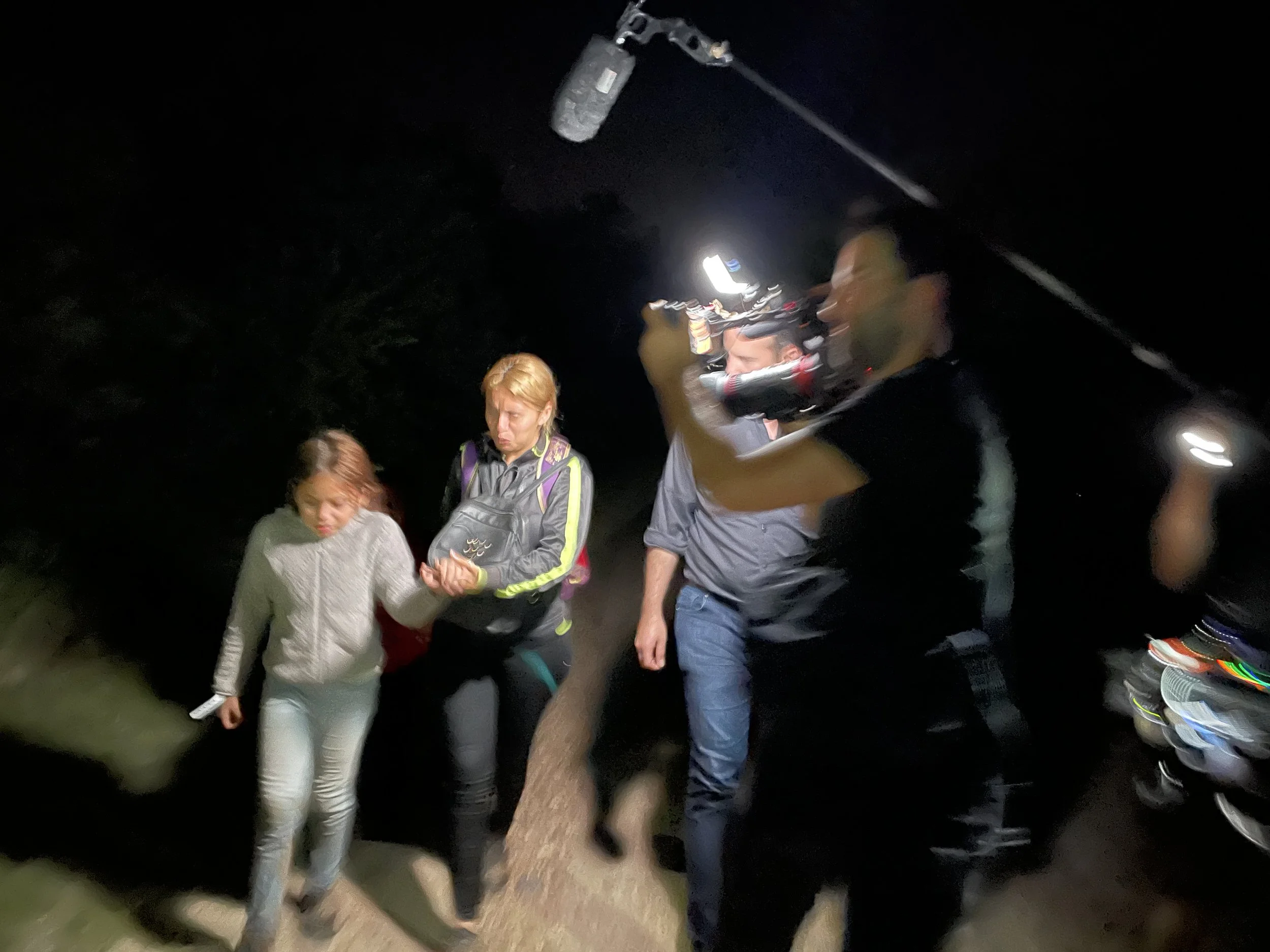Nighttime scene with four people, one supporting a woman who appears to be injured, with a cameraman recording the scene.