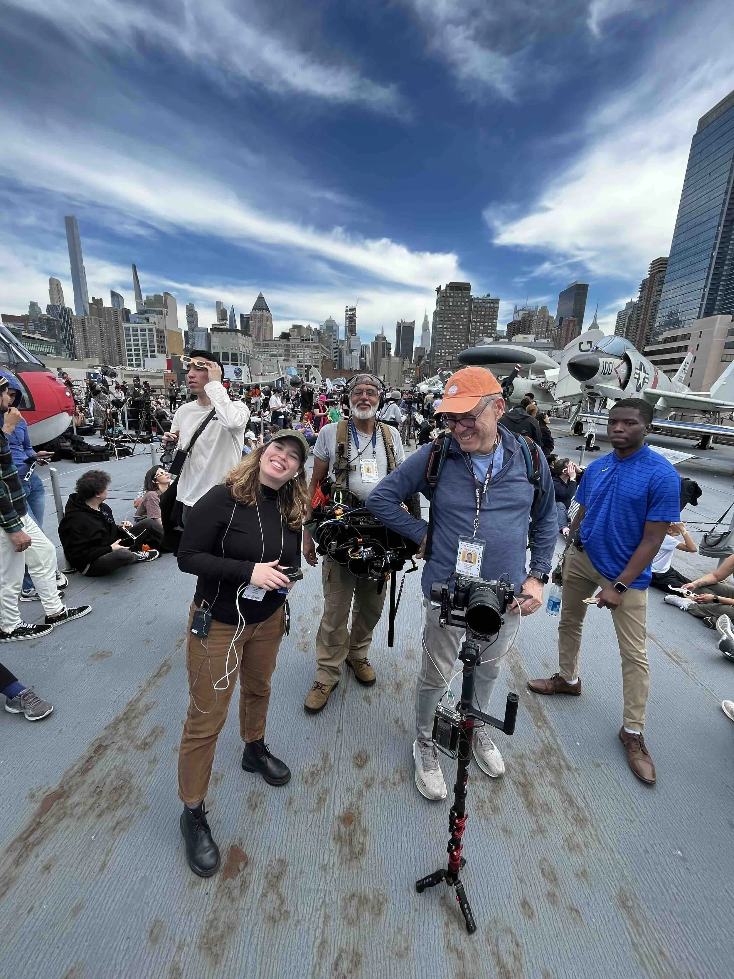 Group of people on an aircraft carrier deck with city skyline in the background, some sitting and others standing, with airplanes and equipment around.