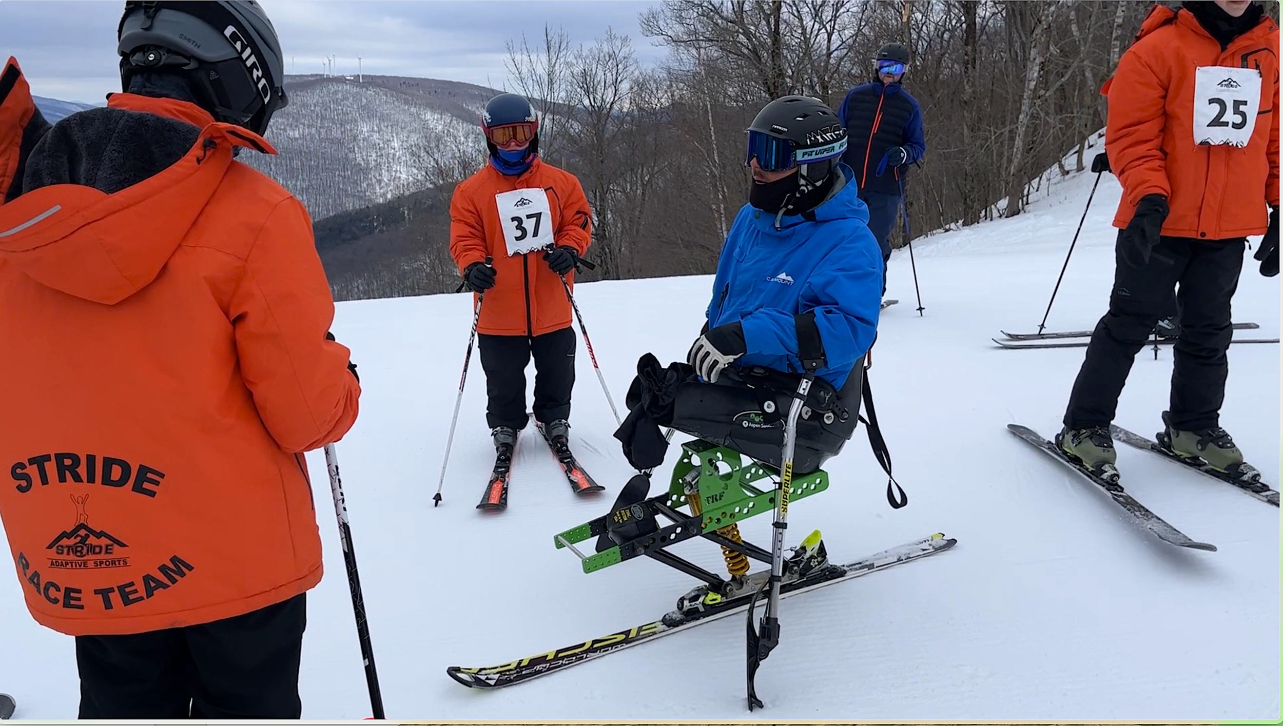Skiers in orange jackets with race bibs on snowy mountain, one skier in a wheelchair equipped for skiing, with a green device on skis, surrounded by other skiers and instructor.