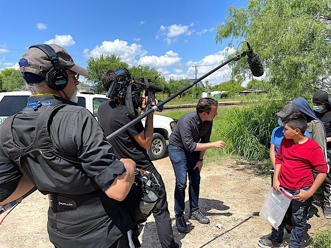 A scene of a film crew interviewing a young boy outdoors near greenery, with a man pointing and speaking to him, while a cameraman films the interaction. Crew members are wearing casual clothing and face masks, and a boom microphone is capturing soun