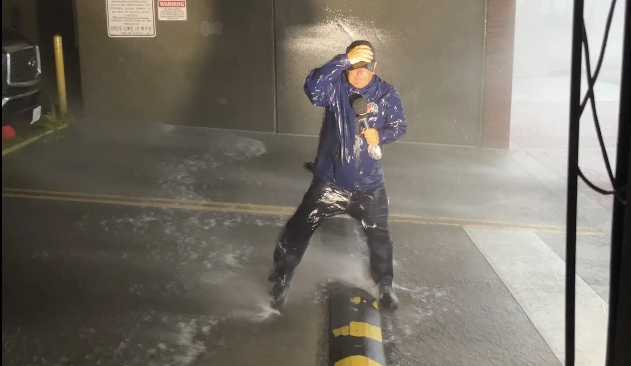 A reporter in a blue raincoat holding a microphone and wiping their face with one hand in a heavy rainstorm, standing on a street with water splashing around.