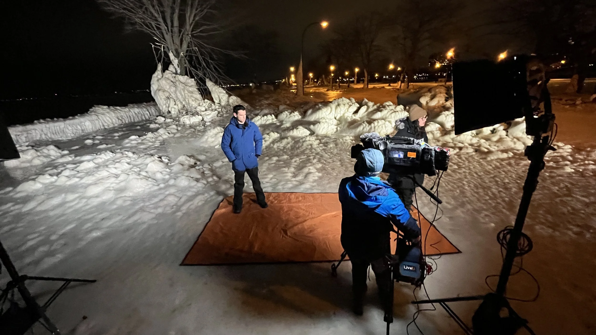 Night outdoor scene with a news crew filming a man standing on a red carpet, surrounded by snow and ice, with streetlights illuminating the background.
