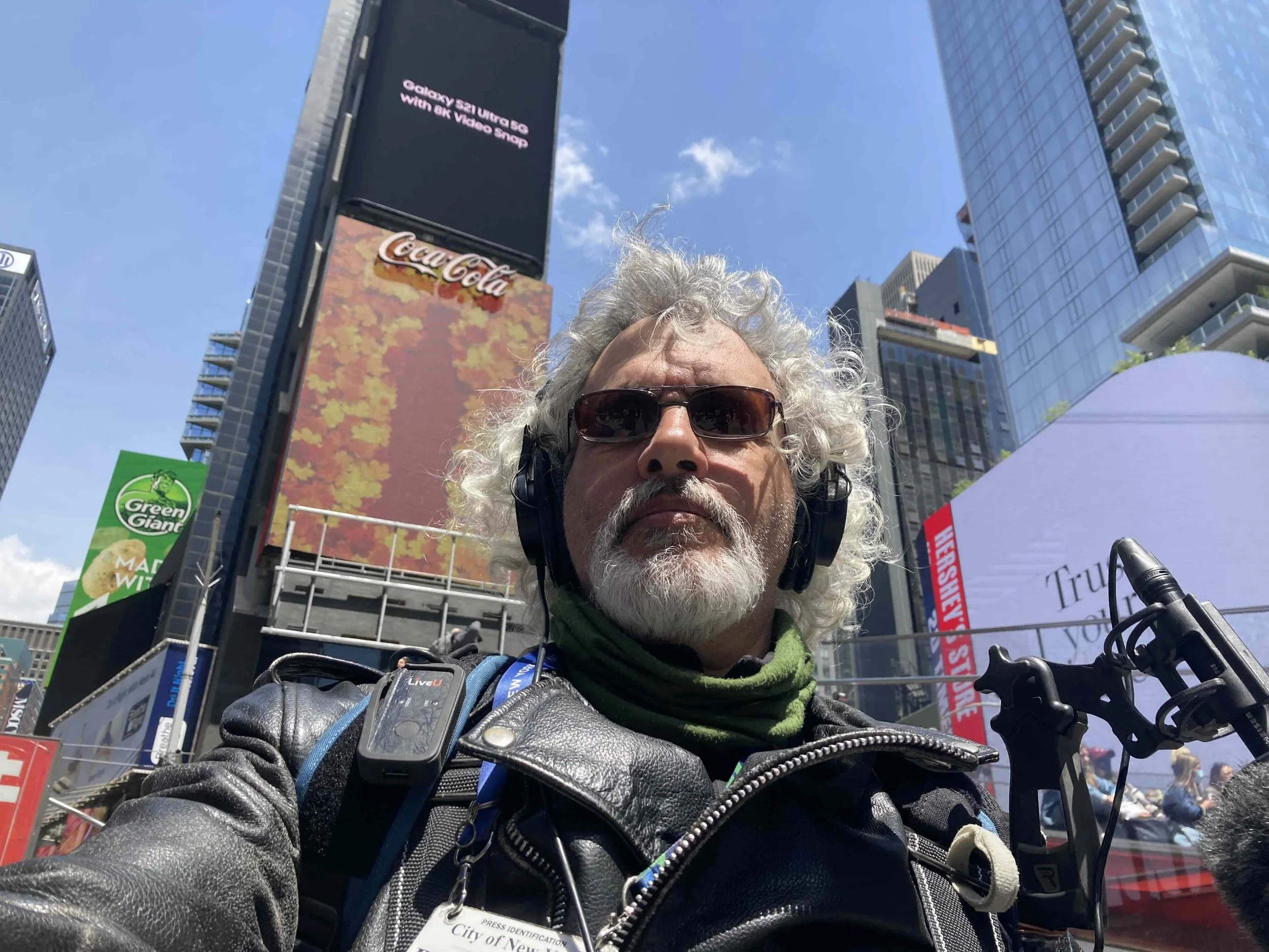 A man with curly gray hair, wearing sunglasses, a leather jacket, and a green scarf, taking a selfie in Times Square with tall buildings, digital billboards, and a crowd in the background.