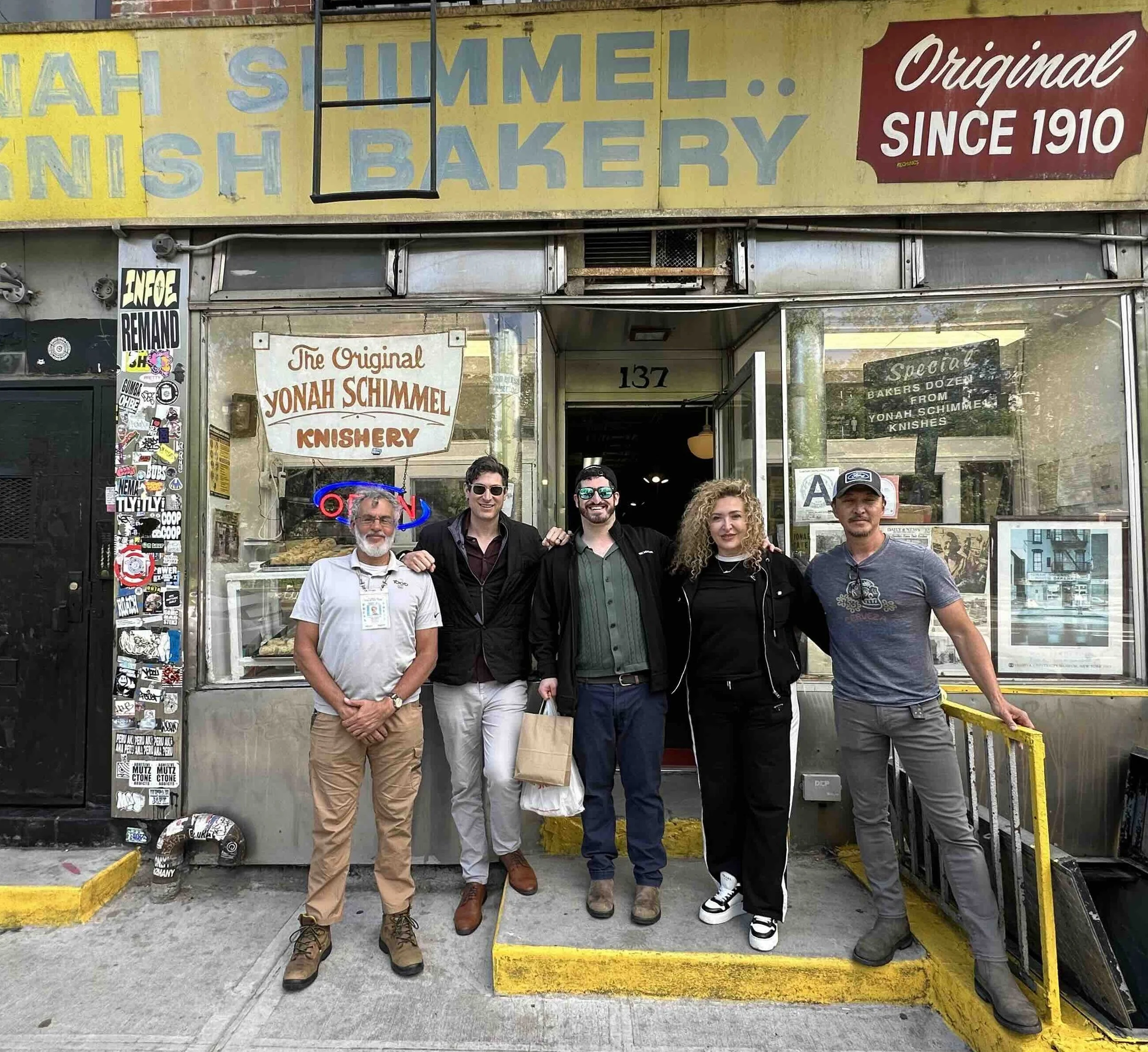 Five people standing in front of Yonah Shimmel Knishery bakery, with signs indicating it has been in operation since 1910. The group is smiling, with some wearing sunglasses and casual attire. The bakery's storefront is decorated with various sticker