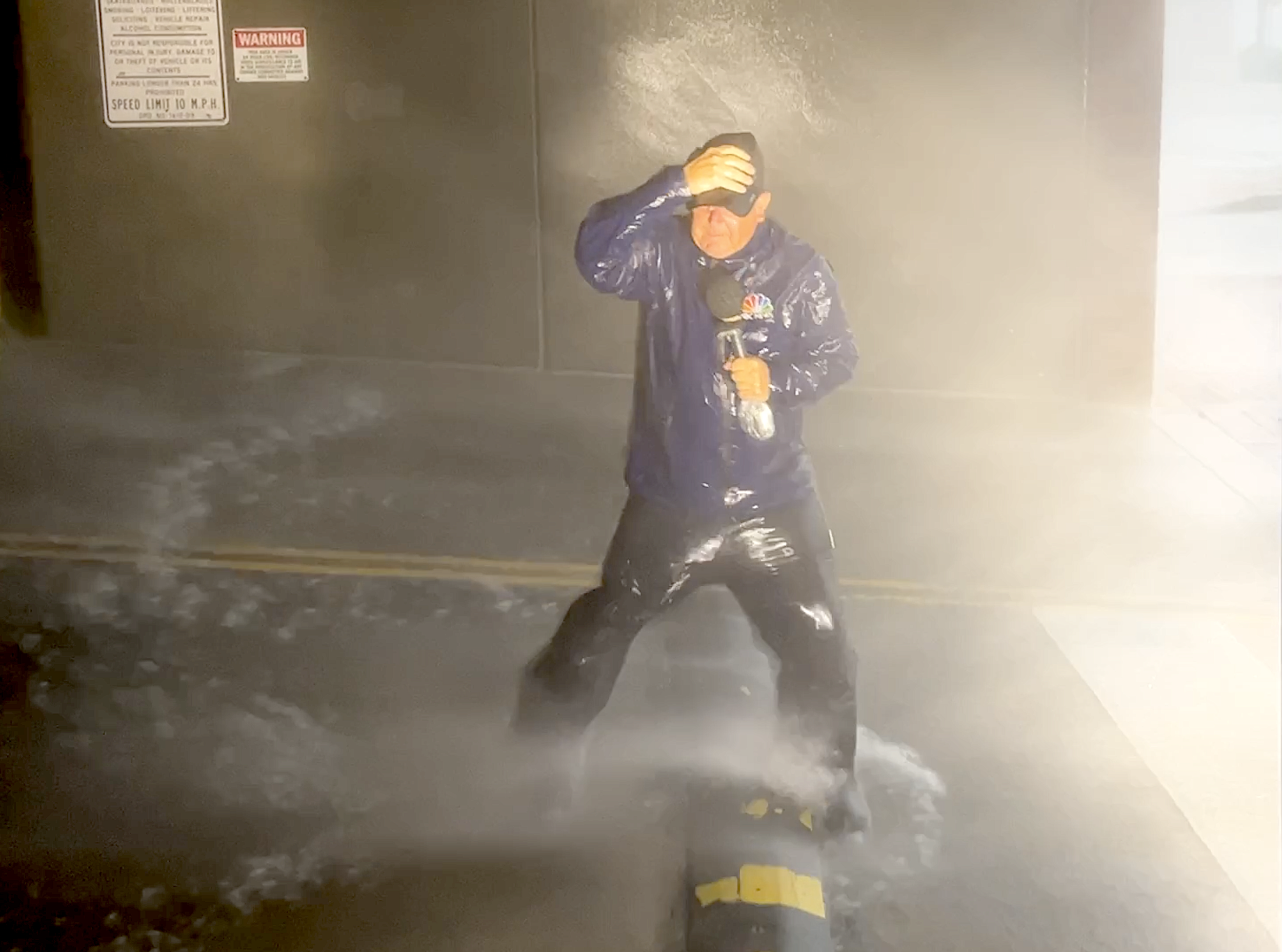 A journalist stands on a black water rescue tube in a flooded street, holding a microphone and shielding his eyes from the rain with his hand.