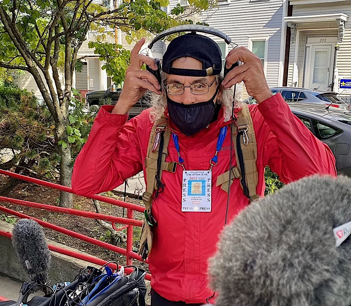 A man with gray hair, glasses, and a black face mask is adjusting headphones. He wears a red jacket and a press badge around his neck. Microphones are directed toward him, and paragraph red railing, trees, house with a white door, and parked cars are