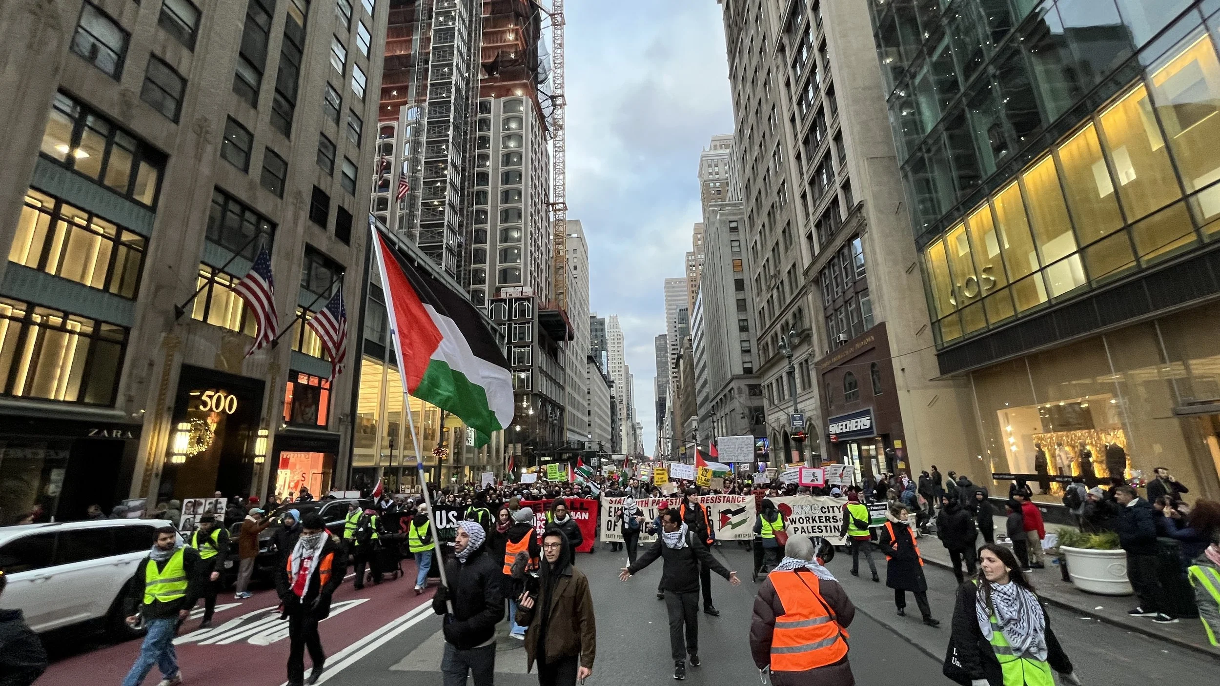 Protest march in a city street with skyscrapers on both sides, people holding signs and flags including the Palestinian flag, some wearing yellow safety vests and scarves, during the daytime.