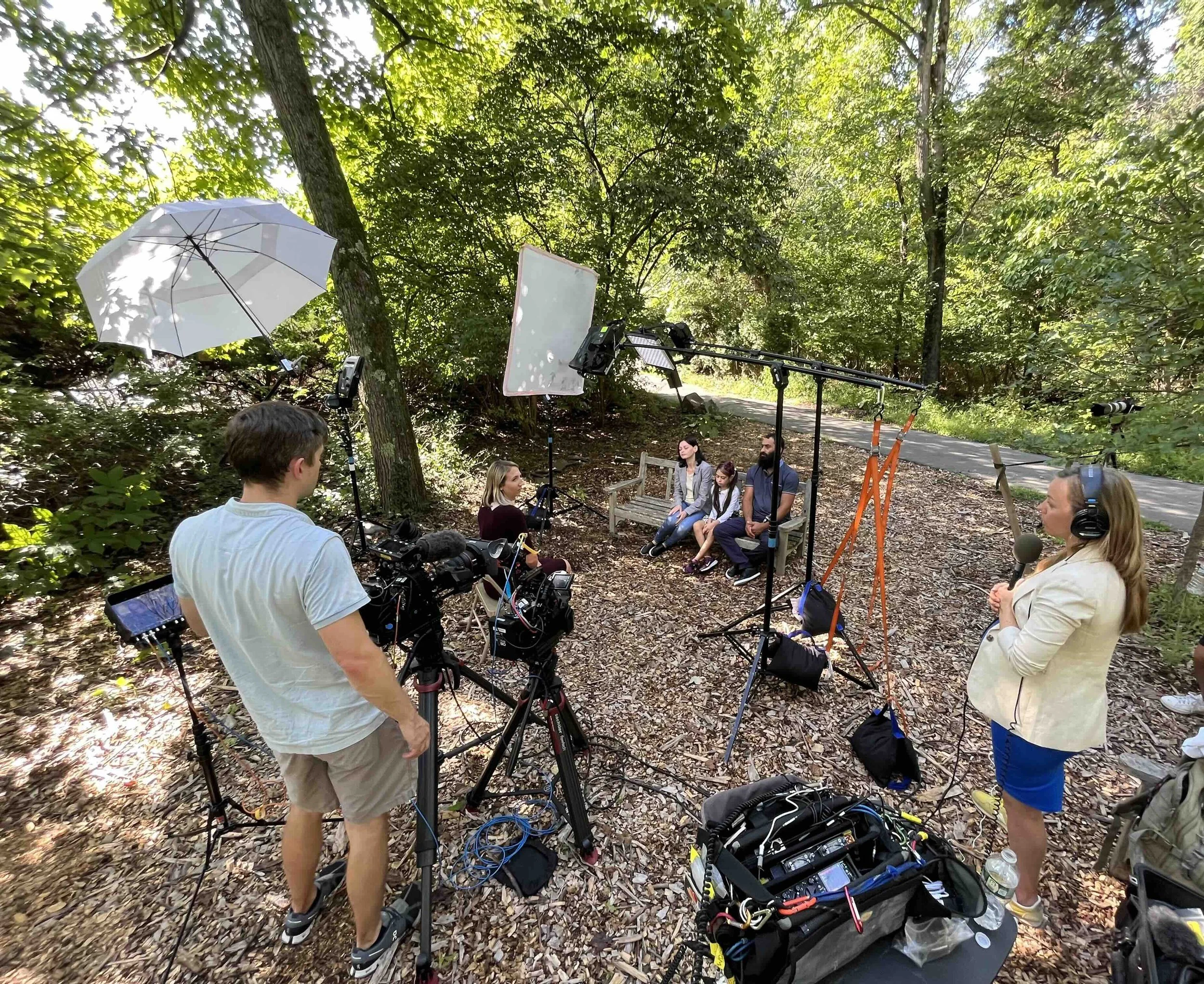 Film crew filming a panel interview with three people seated on a park bench in a wooded outdoor setting, with cameras, lights, and sound equipment.