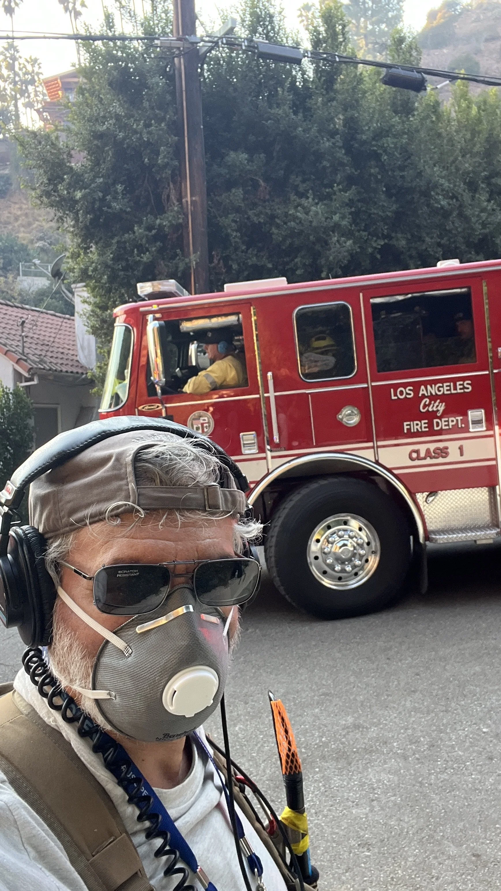 A man wearing sunglasses, a cap, a face mask, and headphones taking a selfie on a street with a red Los Angeles fire truck in the background.