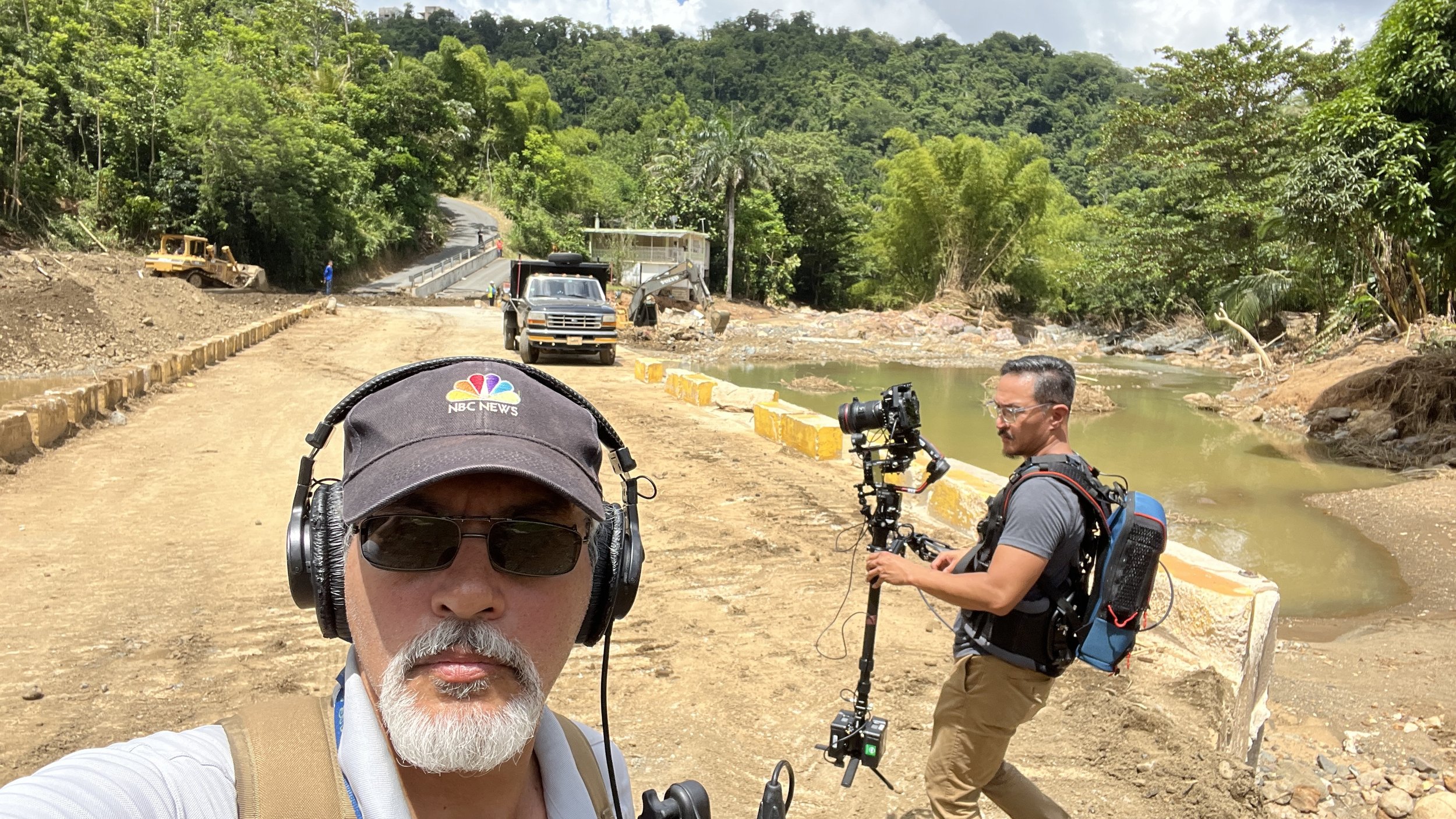 Two men working outdoors on a dirt road near a small body of water with construction equipment and vehicles in the background, surrounded by green trees.