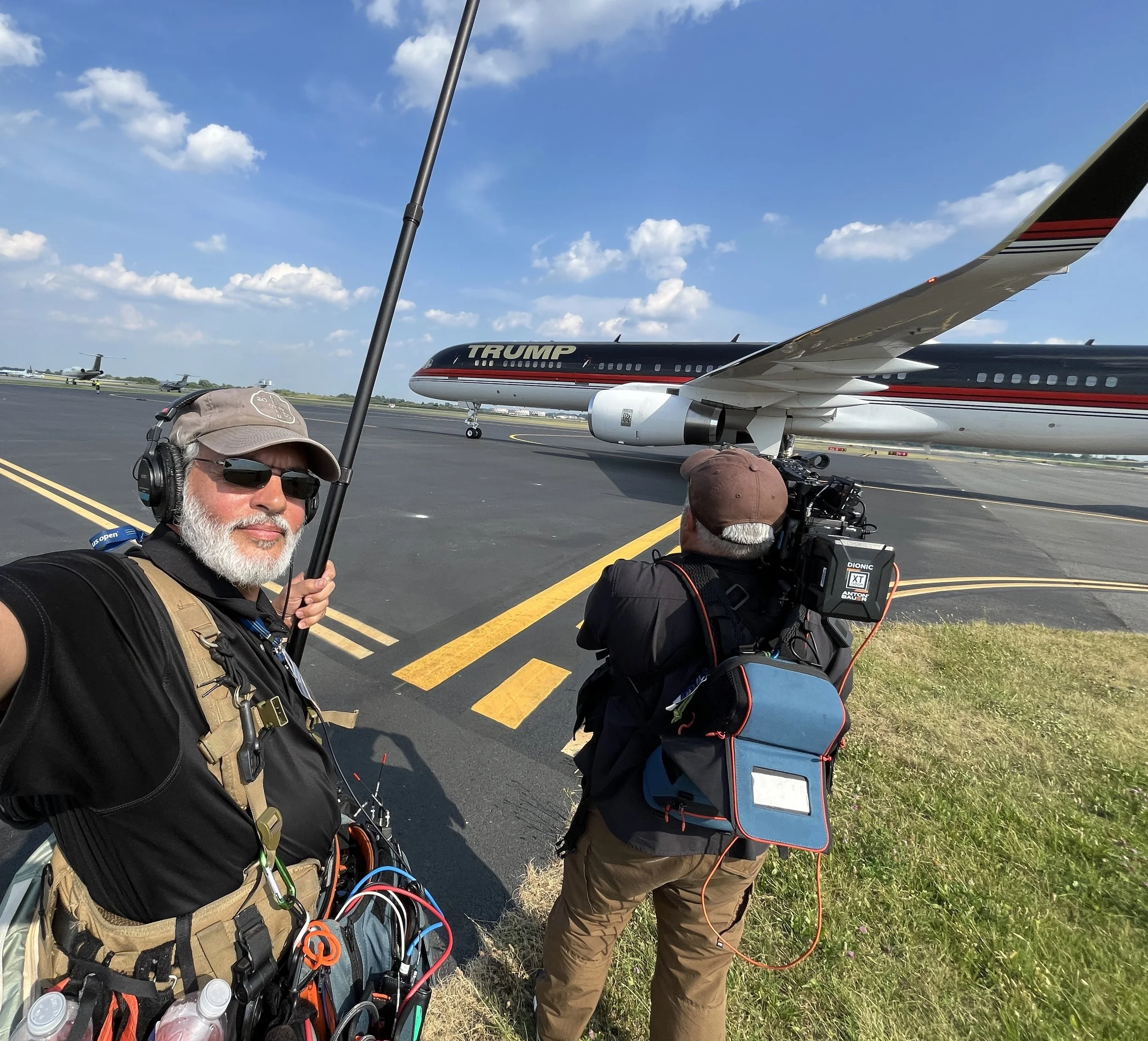 Two men working on an airfield with a custom plane that has 'TRUMP' painted on its side. One man taking a selfie, the other filming the plane. The sky is partly cloudy.