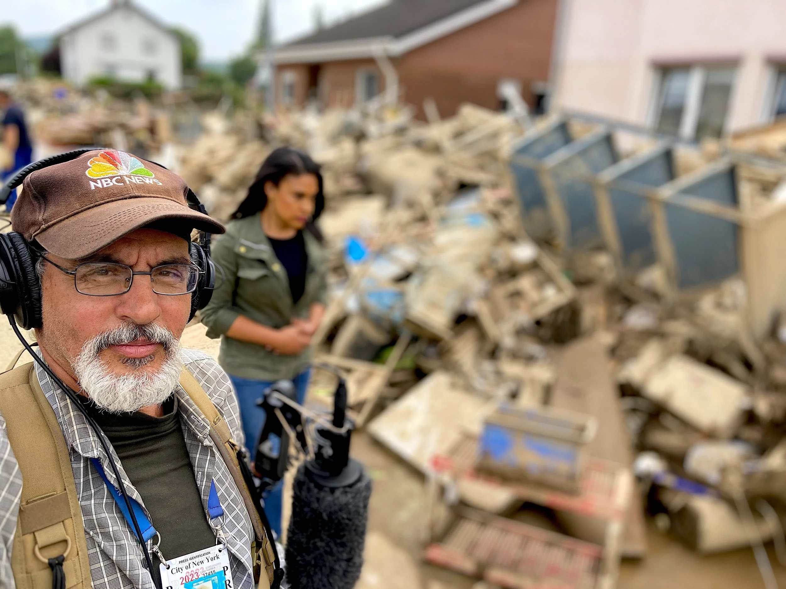 A man with glasses and a beard wearing a NBC News cap and headphones stands in front of extensive debris from destroyed buildings, with a woman in the background amid a scene of destruction.