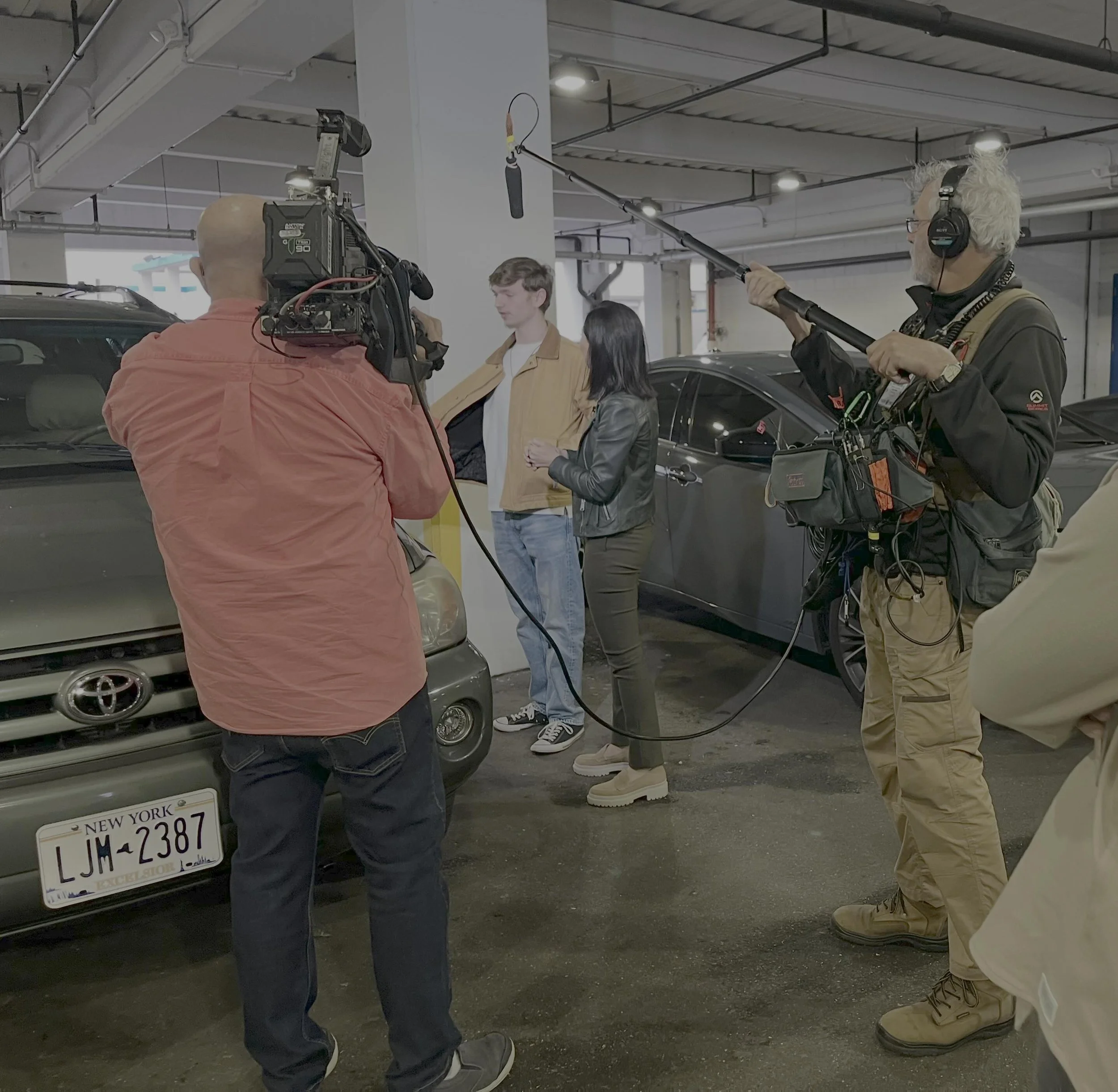 People filming in a parking garage, with two individuals being interviewed and crew members operating cameras and audio equipment.