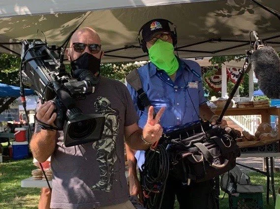 Two men outdoors at a booth, one holding a professional video camera and the other wearing a green face mask and holding a boom microphone, both wearing sunglasses and masks.